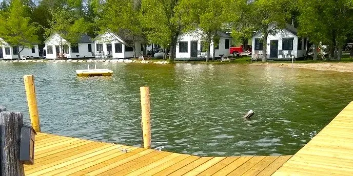 Wooden dock extending over a calm lake with trees and small white houses in the background.