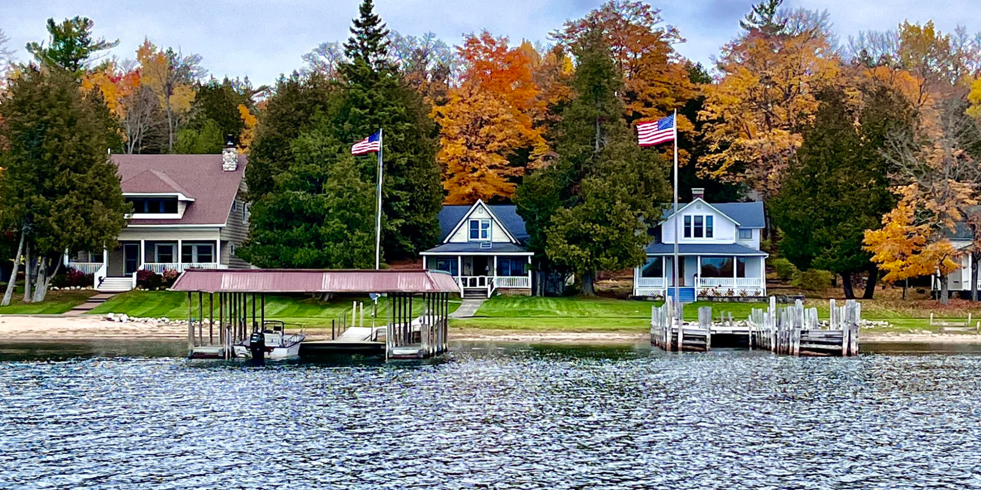 A waterfront scene with houses, trees with autumn foliage, and American flags, with a dock and boat on calm water.