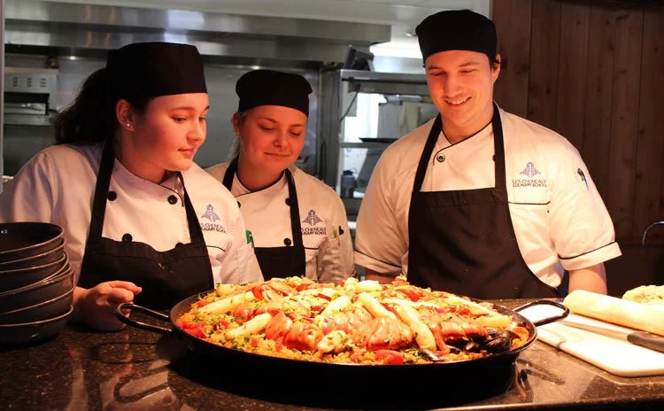 Three chefs in uniform and black hats are standing behind a large pan of cooked seafood paella in a kitchen, smiling and looking at the dish.