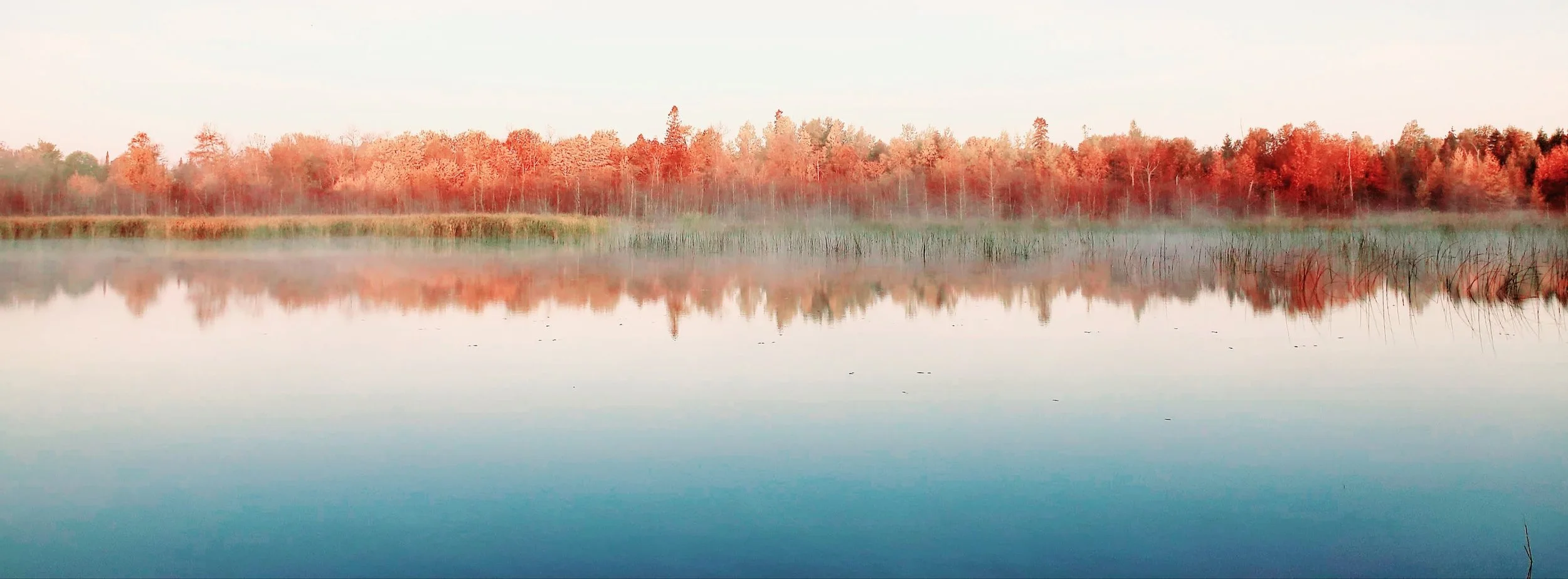 A calm lake with reflections of trees with autumn foliage and some mist on the water surface, during early morning.
