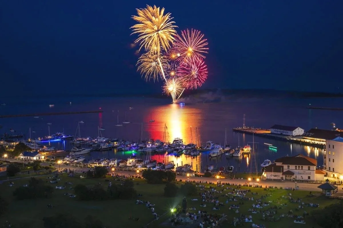 People watching fireworks over a marina at night, with boats docked and reflections on the water.