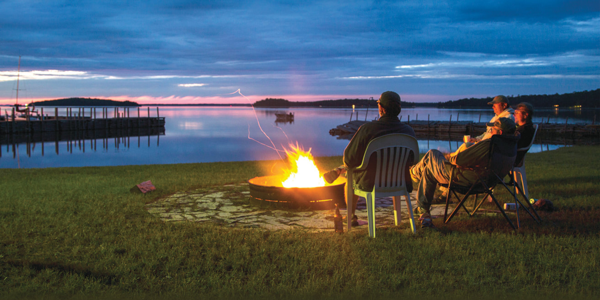 People sitting by a lake at dusk around a firepit, enjoying a campfire with a view of the water, boats, and a distant shoreline under a cloudy sky.