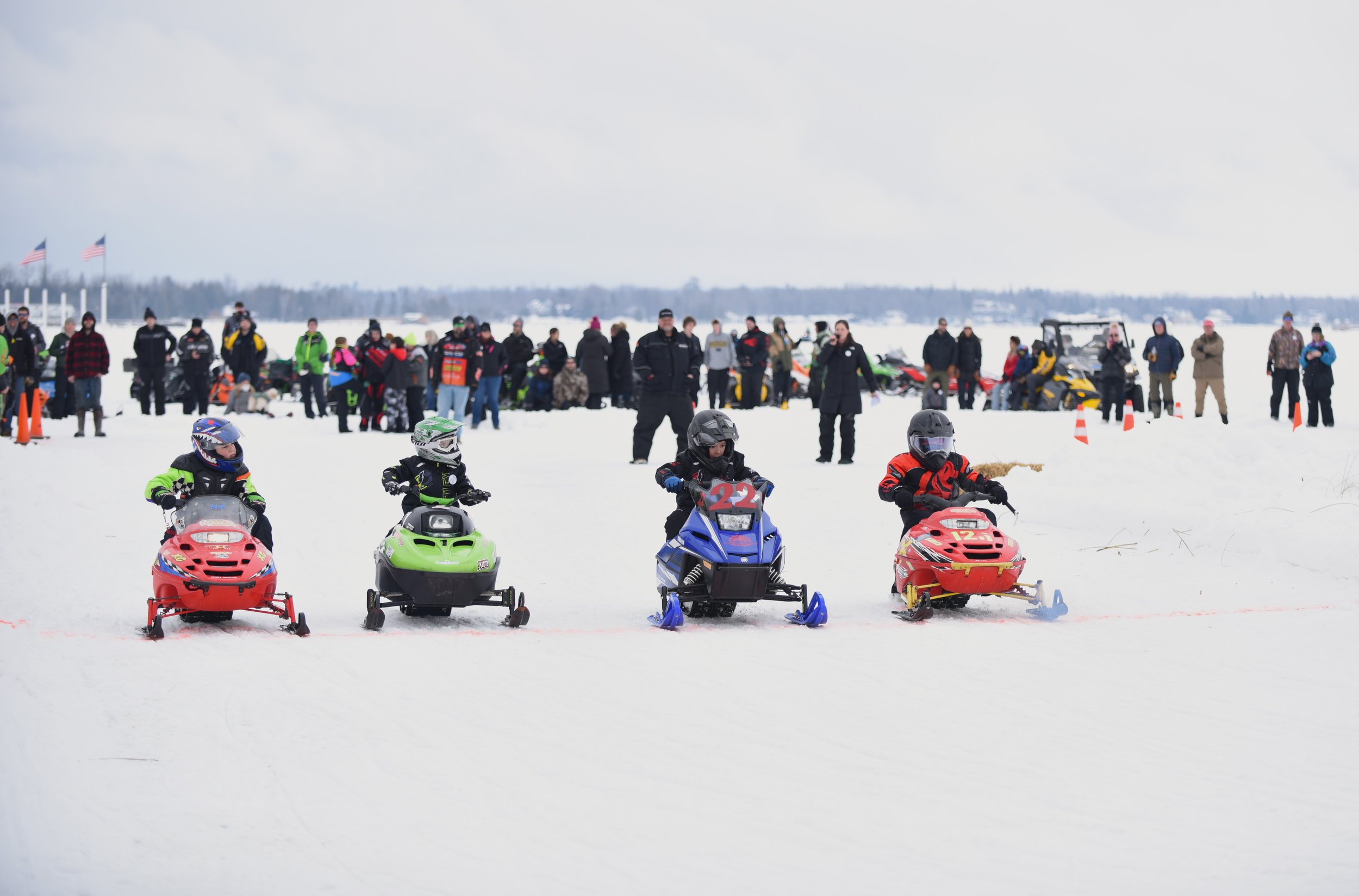 Children riding snowmobiles on a snowy field with numerous spectators and officials watching in the background.
