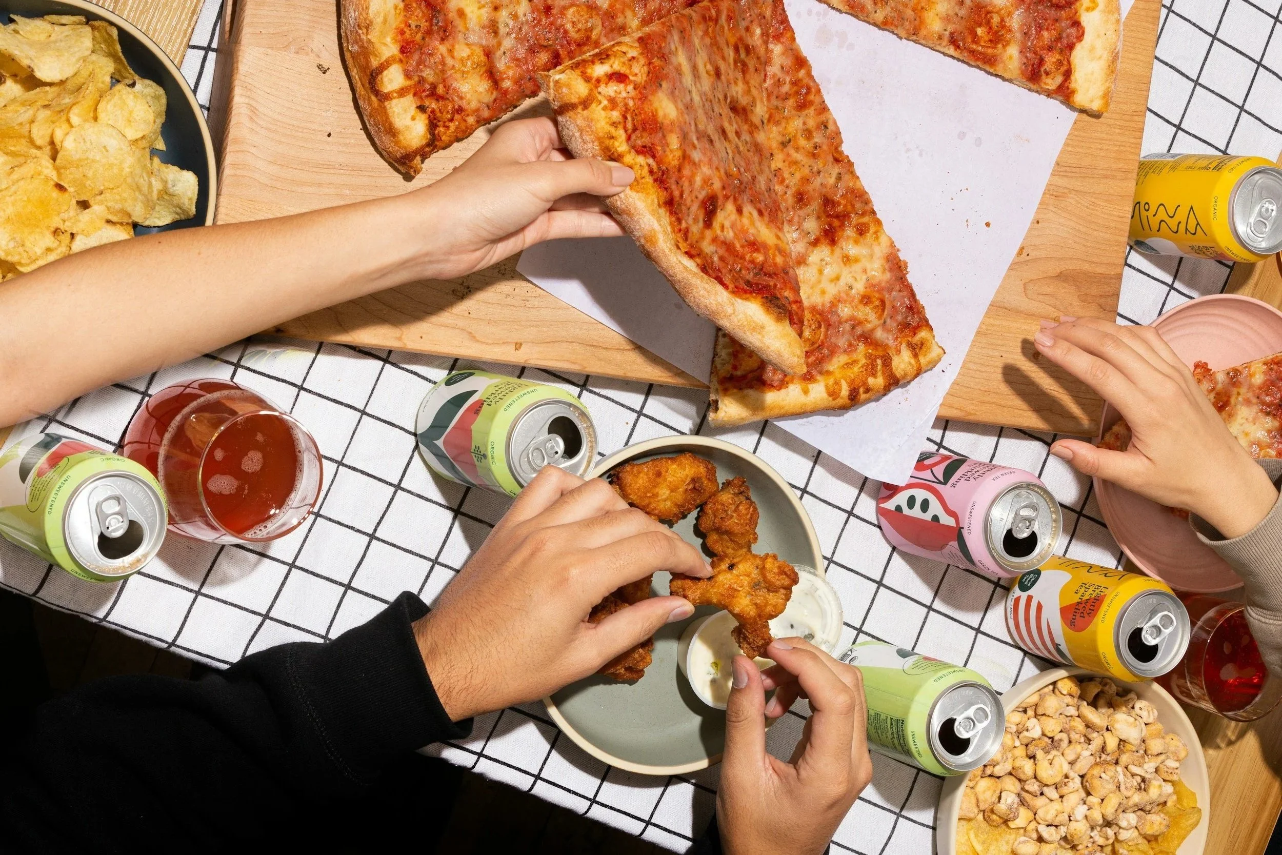 People sharing pizza, chicken wings, chips, and drinks at a casual gathering around a table with a black and white checkered tablecloth.