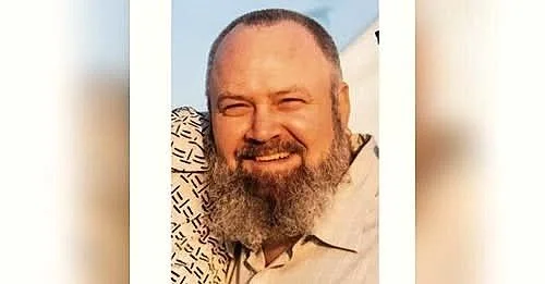 A smiling man with a beard, wearing a light-colored shirt, outdoors with a blue sky in the background.