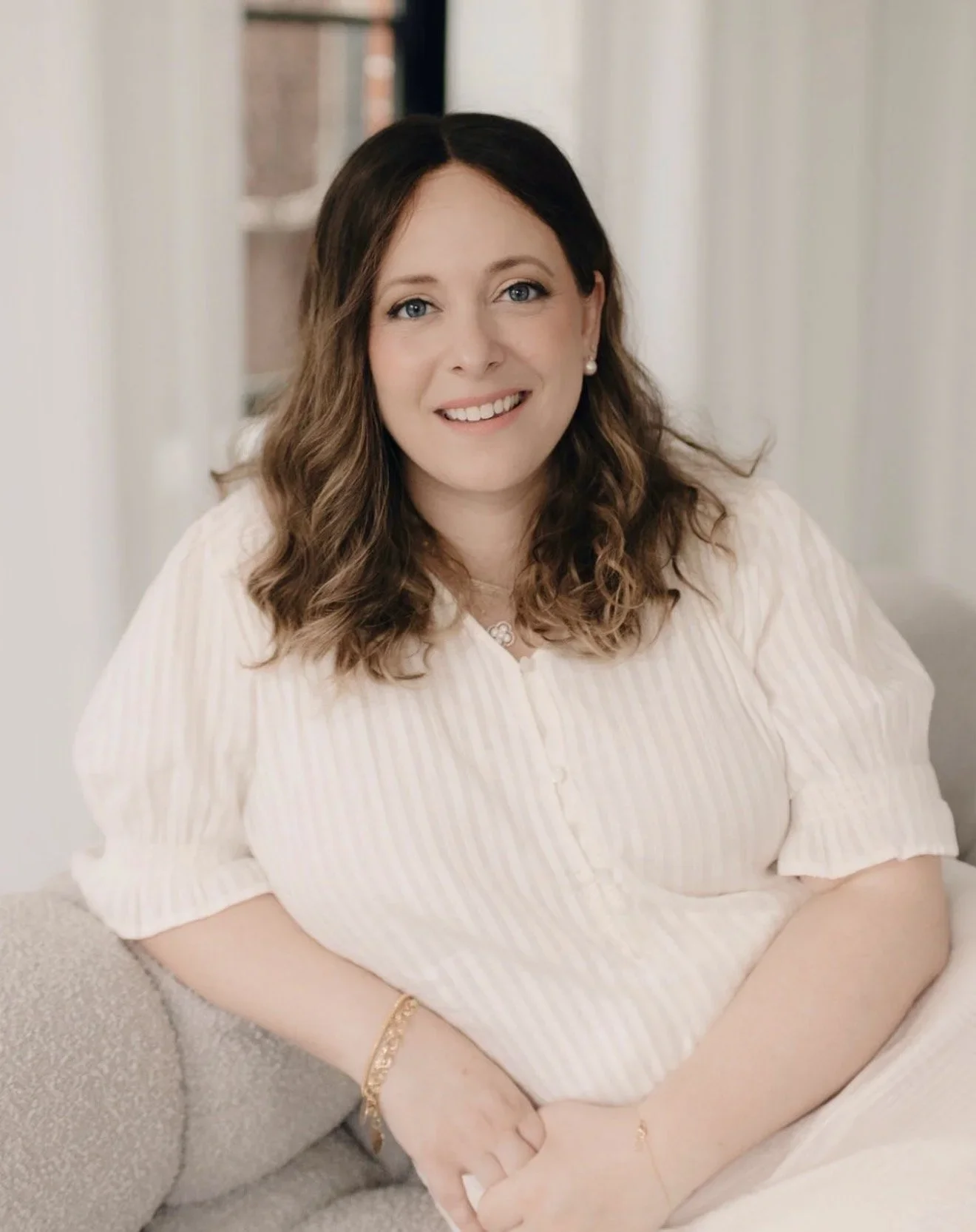 Woman with shoulder-length brown hair and blue eyes, smiling, sitting on a beige couch with a cream wall background.