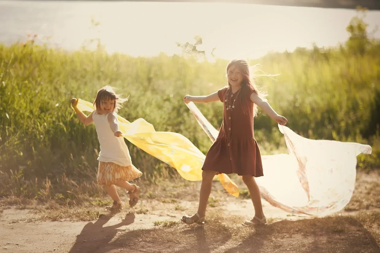 Two young girls playing with colorful scarves outdoors in a grassy field, smiling and enjoying a sunny day.