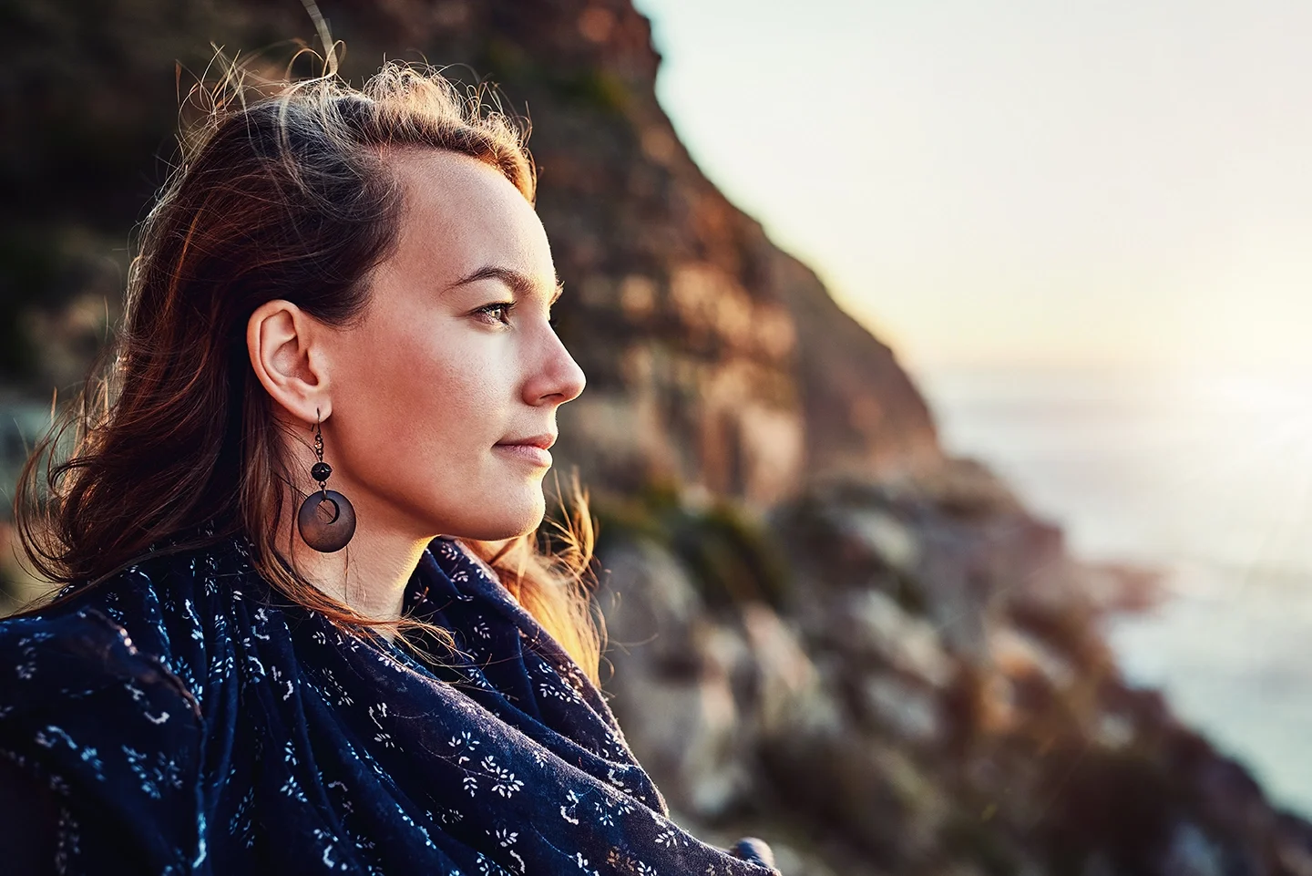 A woman with long wavy hair wearing earrings and a blue patterned scarf, sitting outdoors near a rocky coastline during sunset.