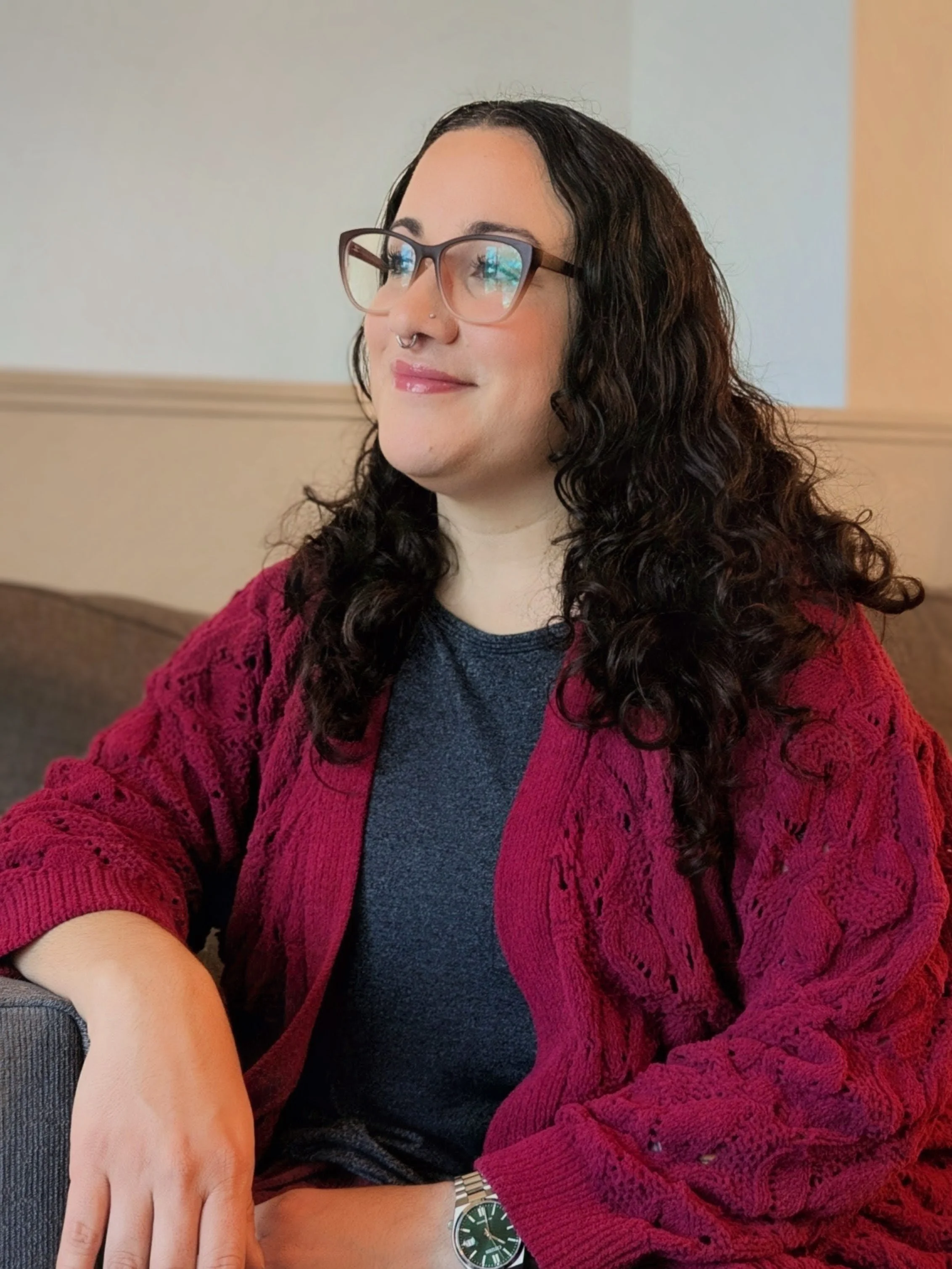 A Florida licensed therapist with long curly dark hair wearing glasses, a nose piercing, a gray top, a red knit cardigan, and a silver watch on her wrist, sitting indoors with a neutral background.