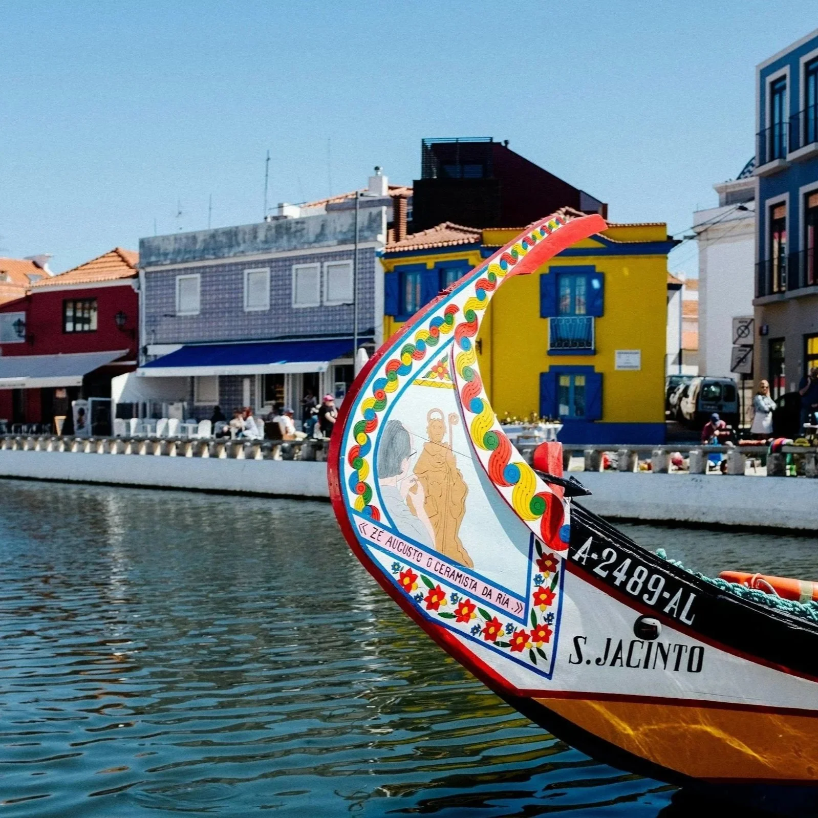 Colorful traditional boat in Aveiro, Portugal, with decorative patterns and images, floating on water with a backdrop of European-style colorful buildings along a street.