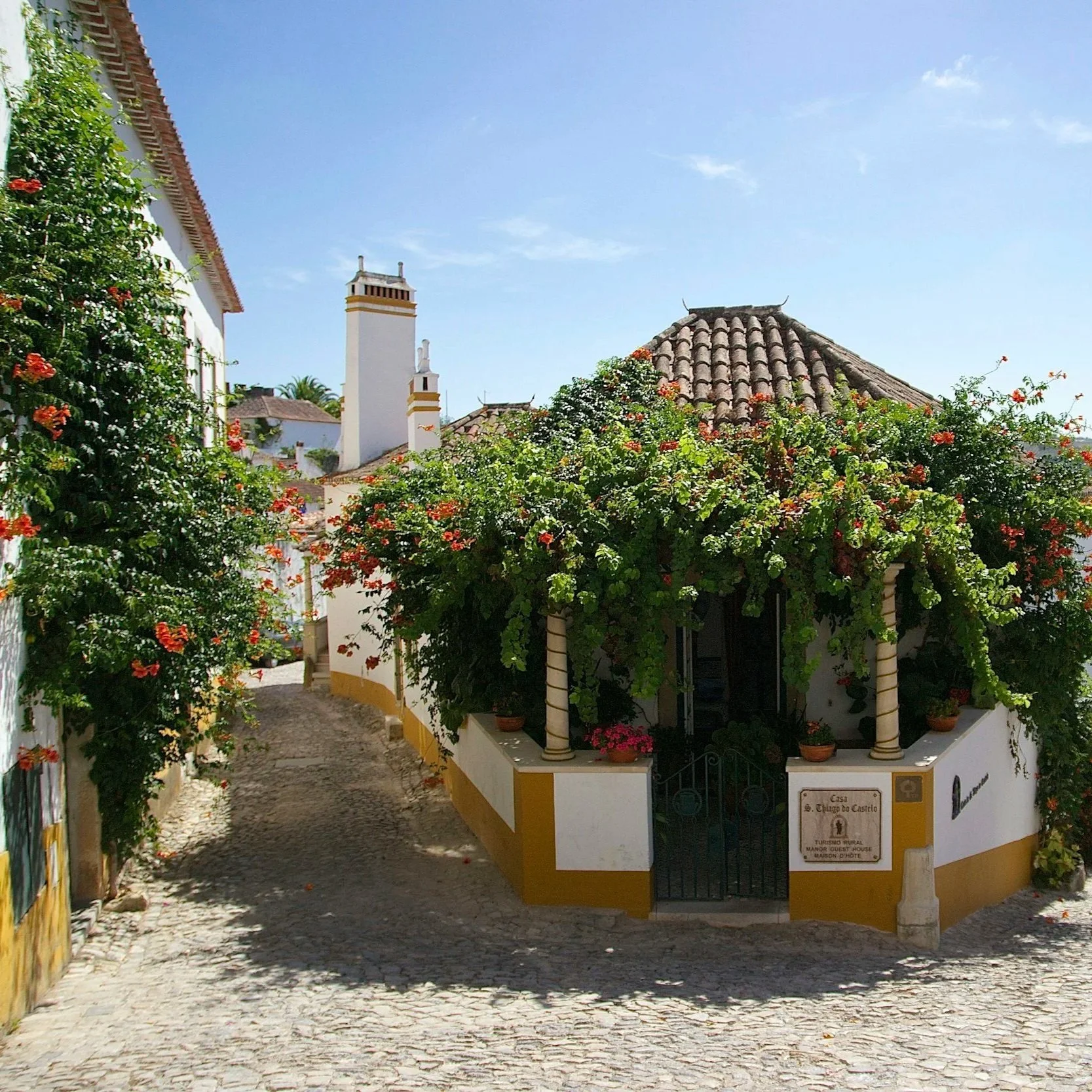 A narrow cobblestone street in Óbidos, Portugal, with white colonial-style buildings adorned with colorful flowers and greenery, under a clear blue sky.