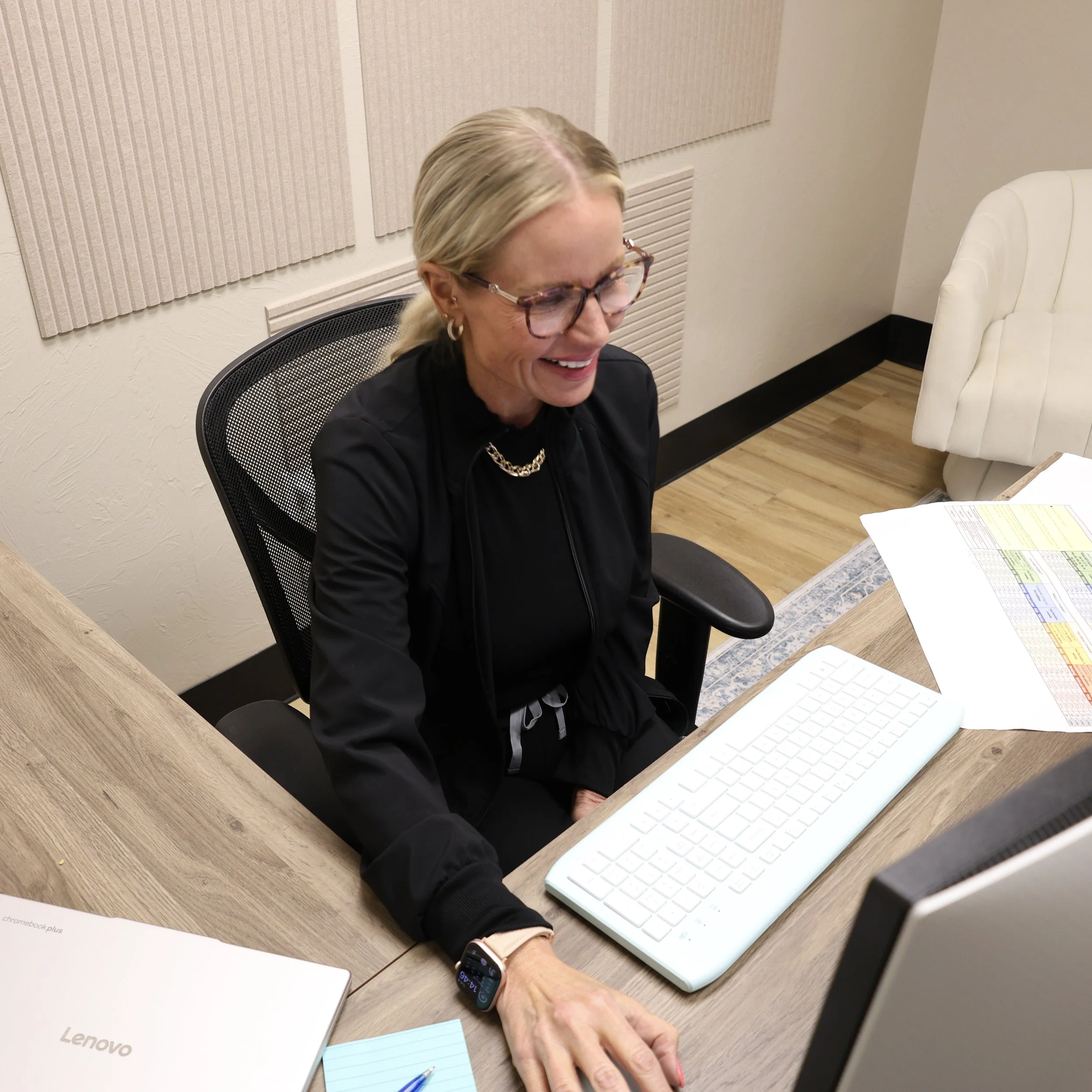 A woman with blonde hair, glasses, and earrings sitting at a desk, smiling while looking at a smartphone. The desk has a white keyboard, a smartphone, notebooks, and papers. There is a white chair and beige acoustic panels on the wall behind her.