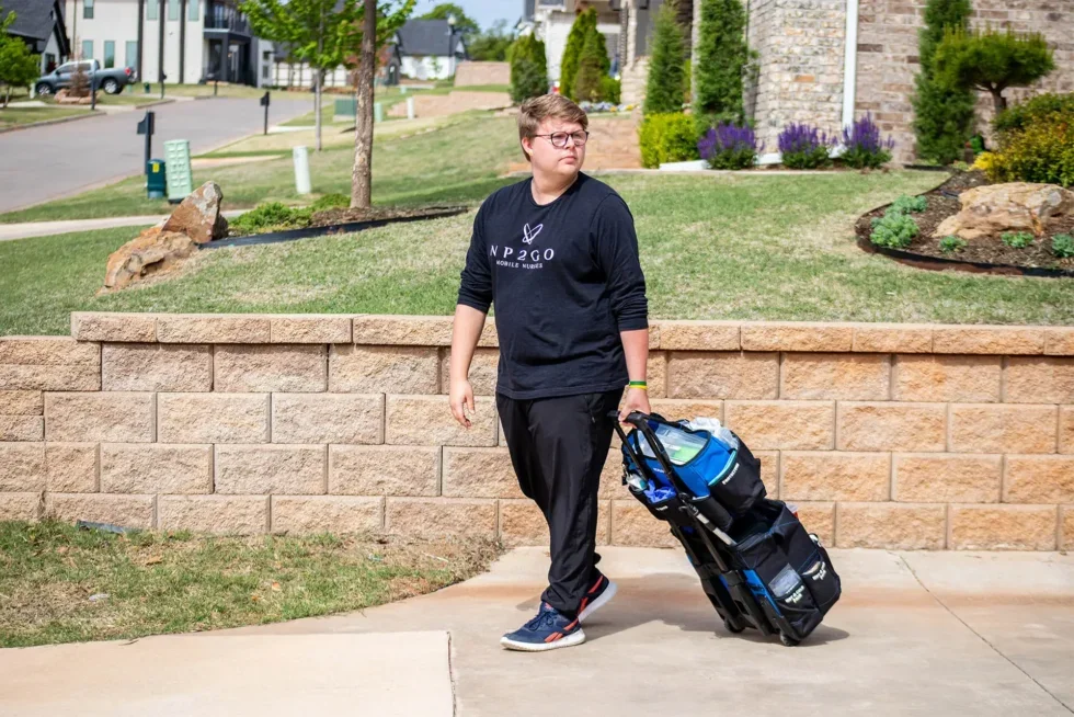 A young person walking on a sidewalk, pulling a rolling bag behind them, with a residential neighborhood in the background.