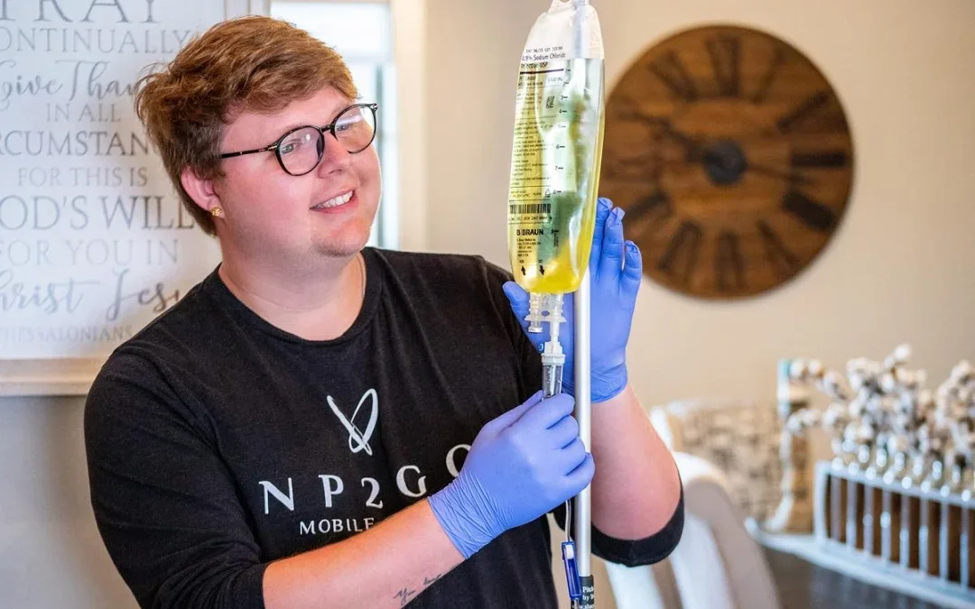 A smiling person wearing glasses and blue gloves holding an IV bag filled with yellow liquid, standing indoors in a room with wall decor, including a large wall clock and framed text.