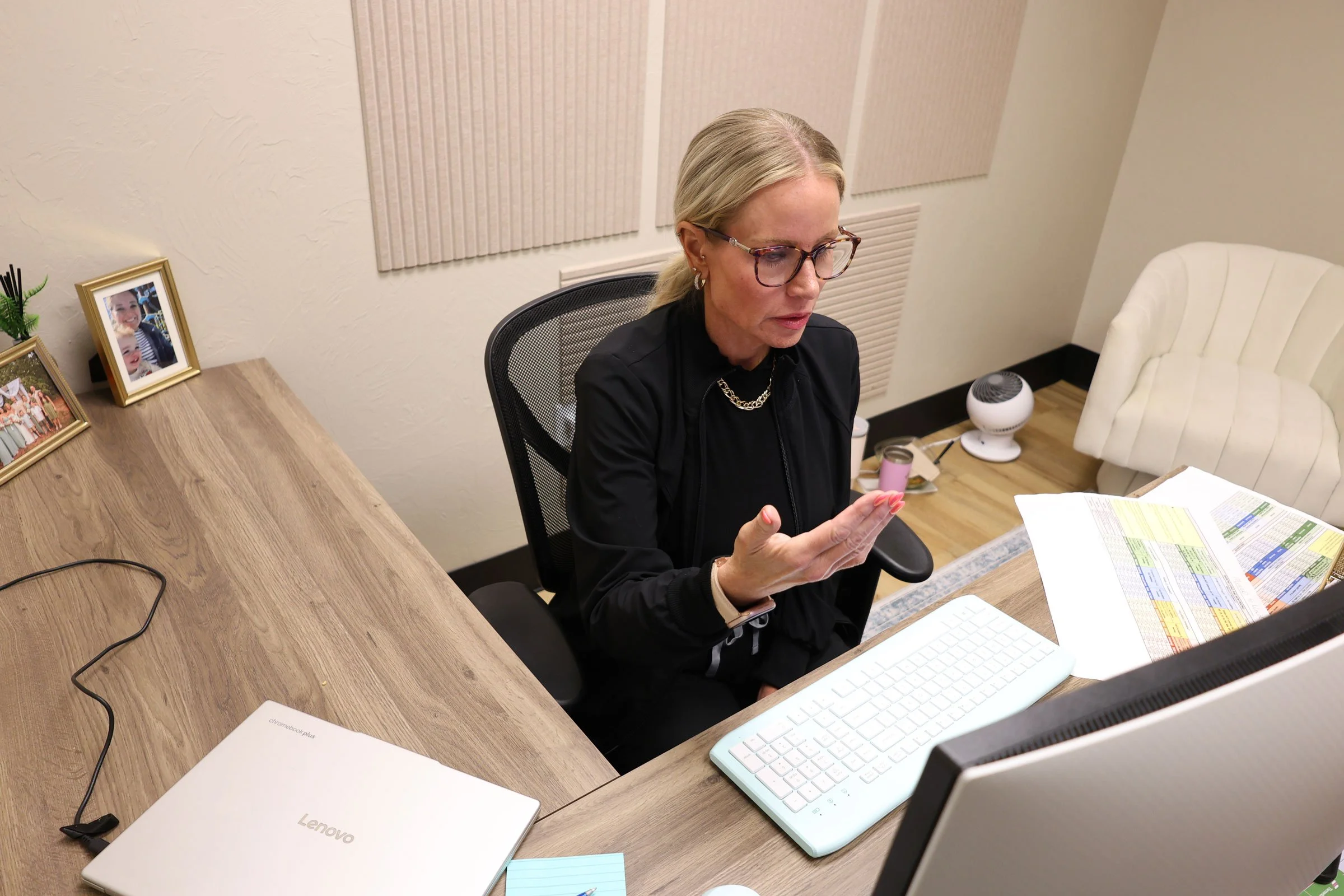 A woman with blonde hair and glasses sitting at a desk, looking at her phone. The desk has a closed white Lenovo laptop, a white keyboard, papers, and a computer monitor. The background features soundproofing panels and framed photos on a side table.