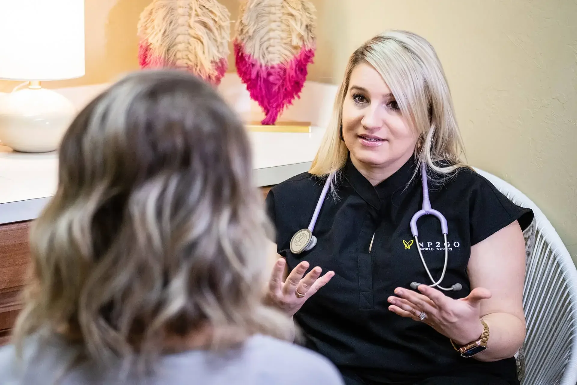 A blonde woman, likely a nurse or healthcare professional, is speaking to a young girl with curly hair in what appears to be a clinical or healing setting. The woman wears a black uniform with a stethoscope around her neck and a badge that says "Noble Nurse" and "2020." She has a gentle, engaged expression, and the environment has warm lighting with decorative pink-and-orange feathers in the background.
