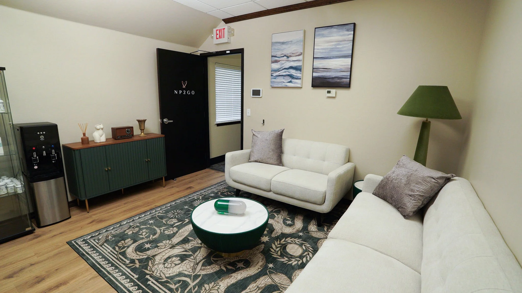A cozy living room with a cream-colored sofa, a green floor lamp, and wall art depicting abstract landscapes. There is a round coffee table with a decorative centerpiece, a patterned area rug, a water dispenser, and a green cabinet with decorative items on top. The room has wood flooring, a window with blinds, and a black door with white text and symbols.