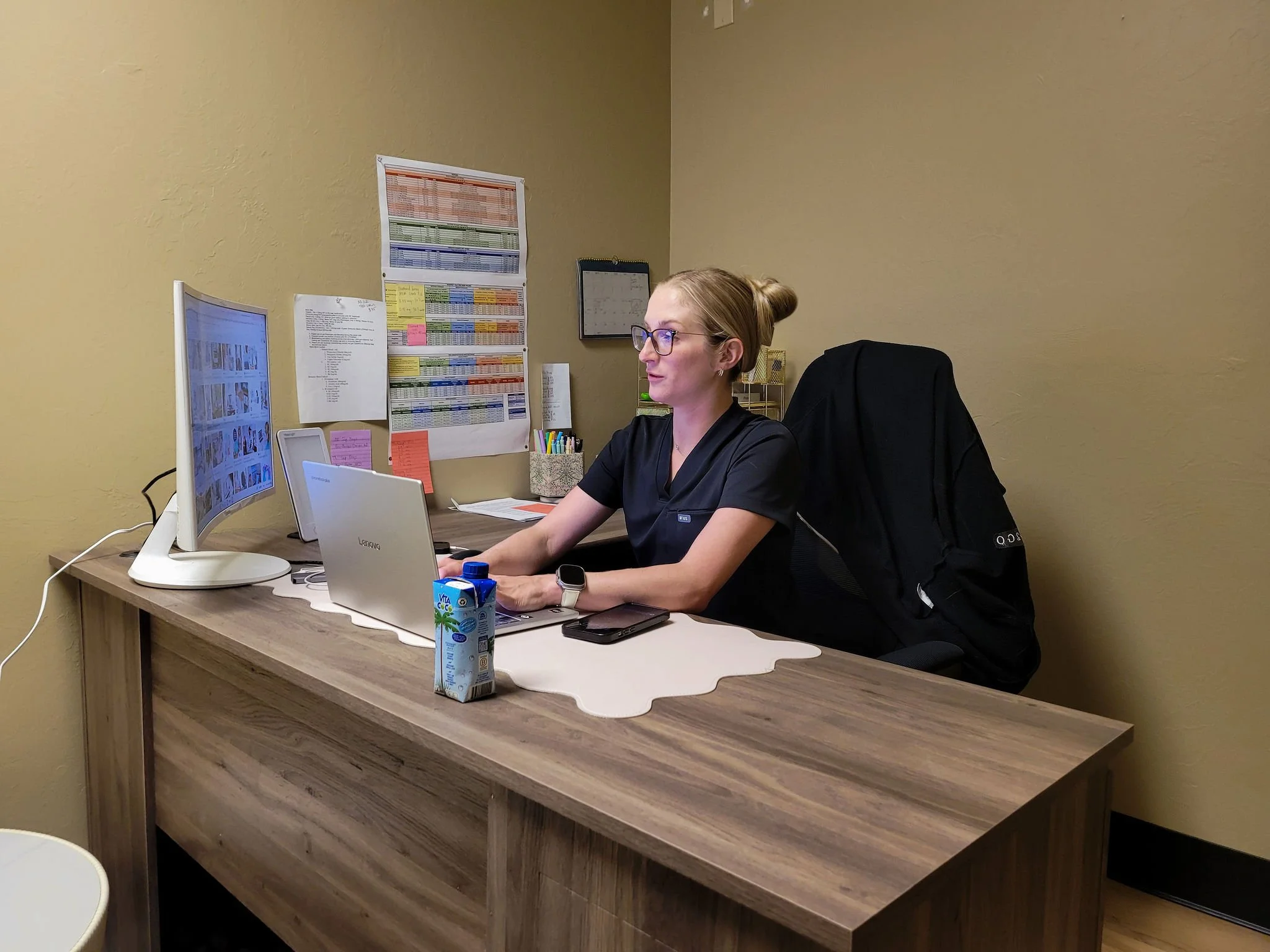 Woman working at a desk in an office, with a computer monitor, laptop, and office supplies, against beige walls decorated with charts and notes.