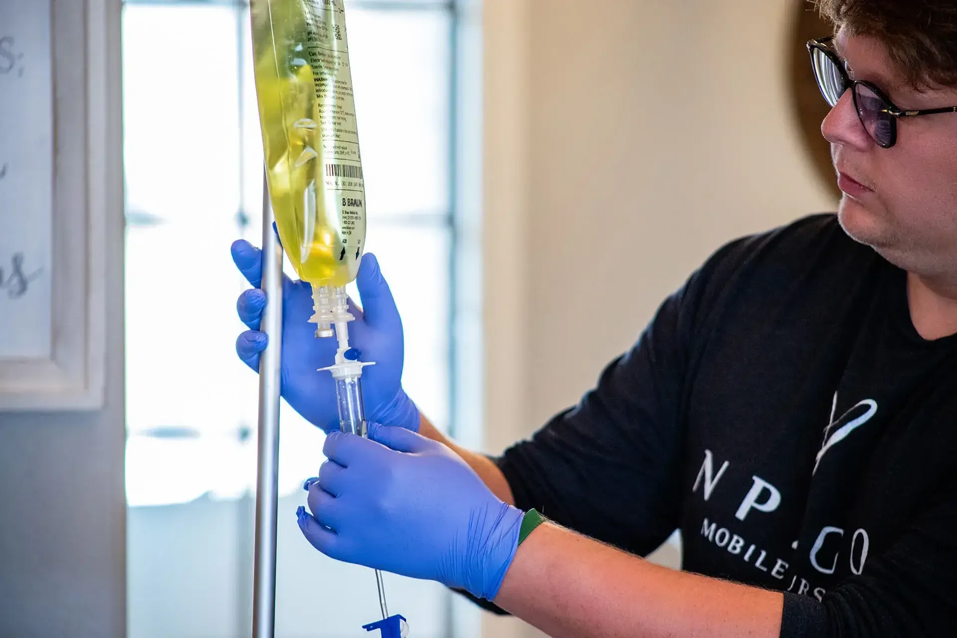 A person wearing glasses and blue gloves preparing a syringe with a yellow liquid, likely a medical fluid, in a clinical setting.
