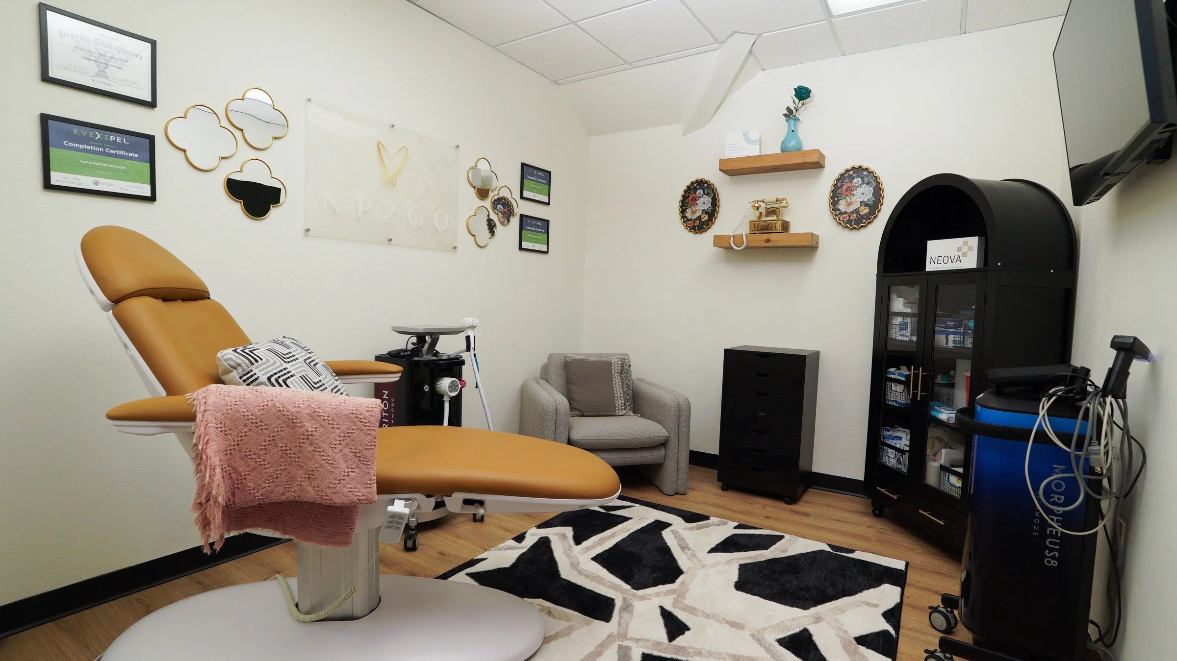 Medical examination room with a reclining chair, two framed certificates, small mirror, decorative wall hanging, small TV, blue vase with flower on shelf, vintage phone, gray armchair, black storage cabinet, and medical equipment including a nebulizer and blood pressure monitor.