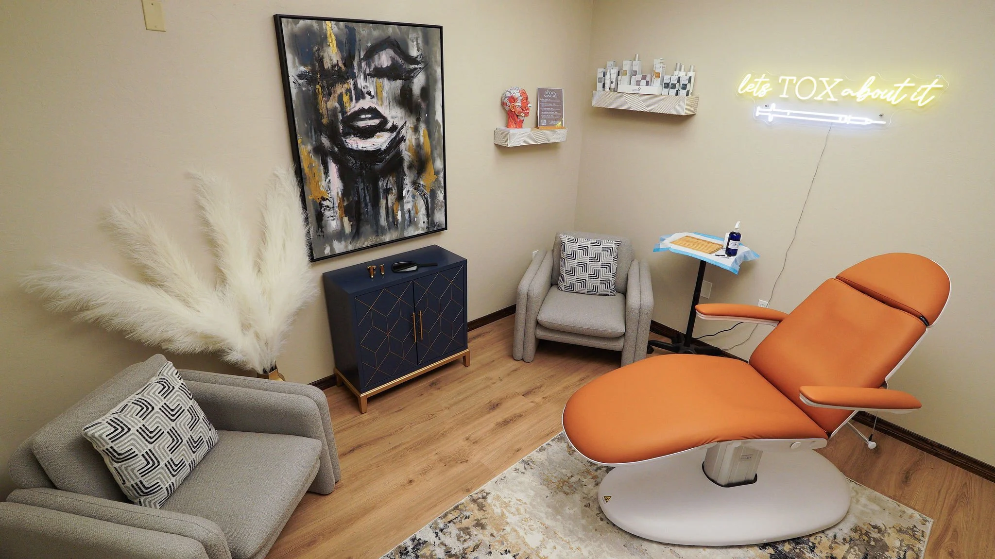A medical examination room with an orange examination chair, a small sofa with black and white patterned pillows, a blue cabinet, wall art, a small armchair, and a neon sign that reads 'let's TOX about it'.