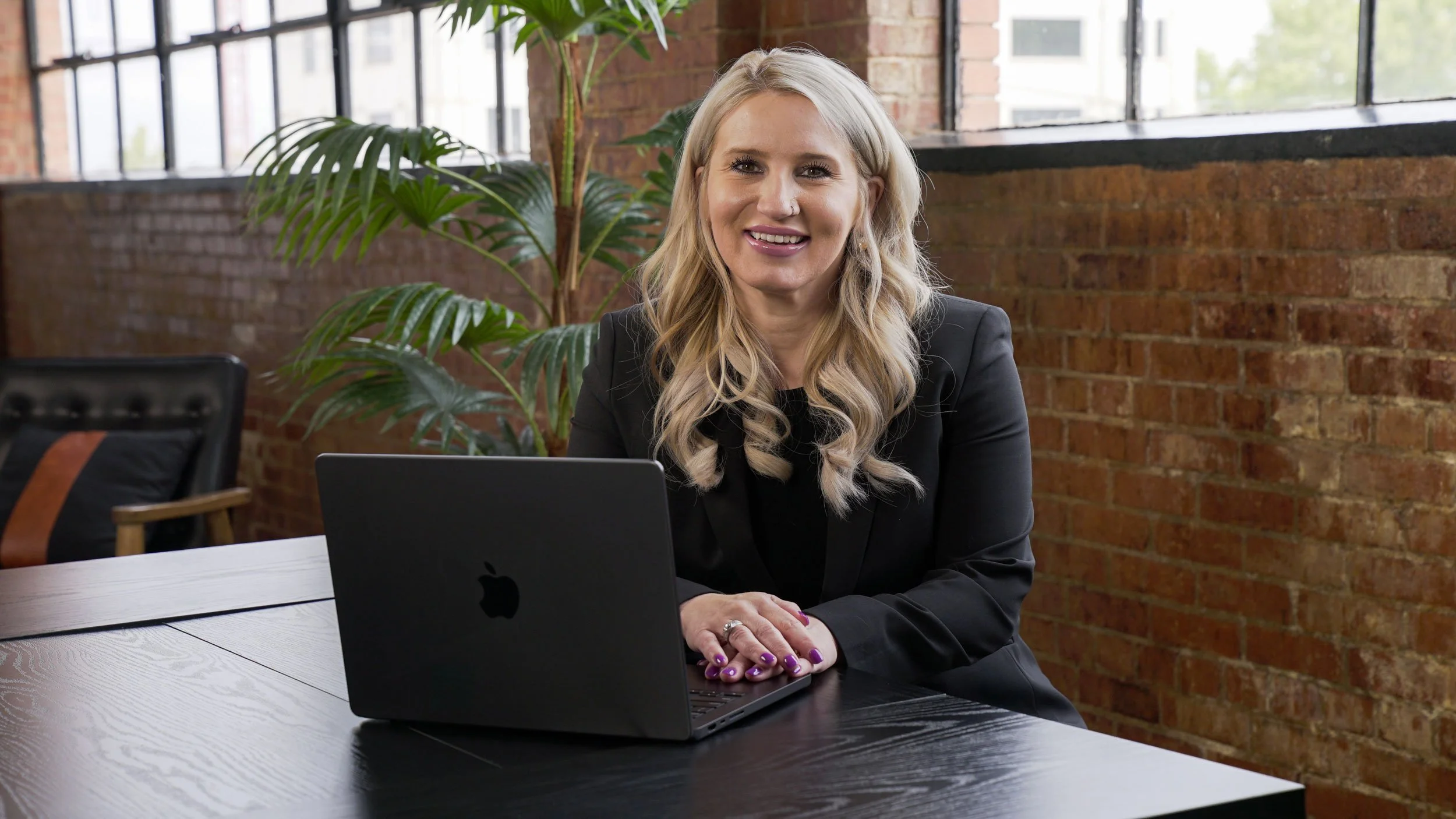 A woman with blonde hair, wearing a black blazer, smiling at the camera, sitting at a desk with a laptop in an office with brick walls and large windows, with green plants in the background.