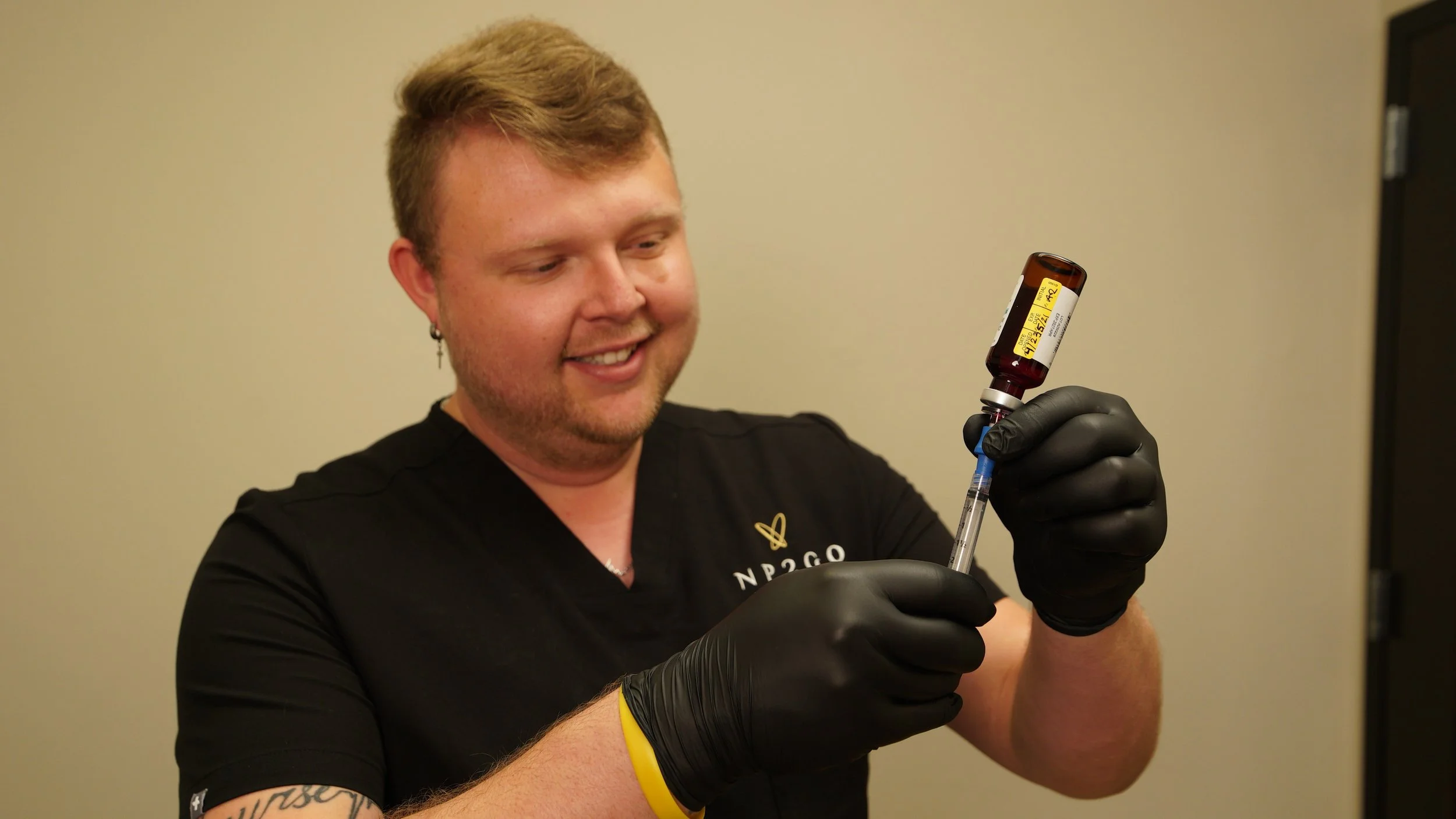 A young man with short blond hair wearing black gloves, a black shirt with a logo, and a yellow wristband, smiling as he prepares a syringe with medication from a brown vial.