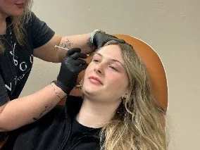 A woman is lying back with her eyes closed while a makeup artist applies makeup to her face in a studio setting.