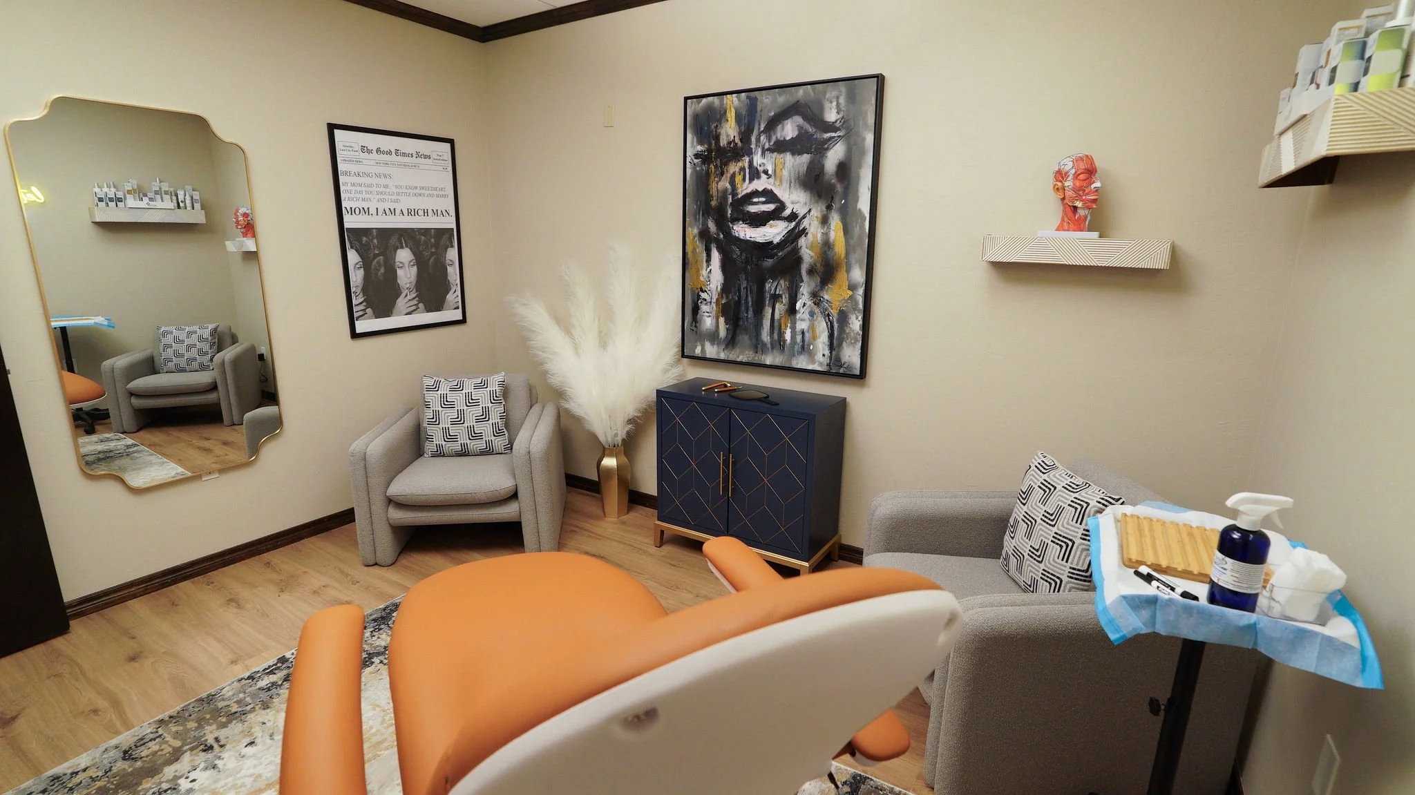 A small waiting room with two gray armchairs, a navy blue cabinet with gold accents, a large abstract portrait of a woman on the wall, shelves with decorative objects, and a mirror reflecting another chair. There are medical supplies on a tray in the foreground.