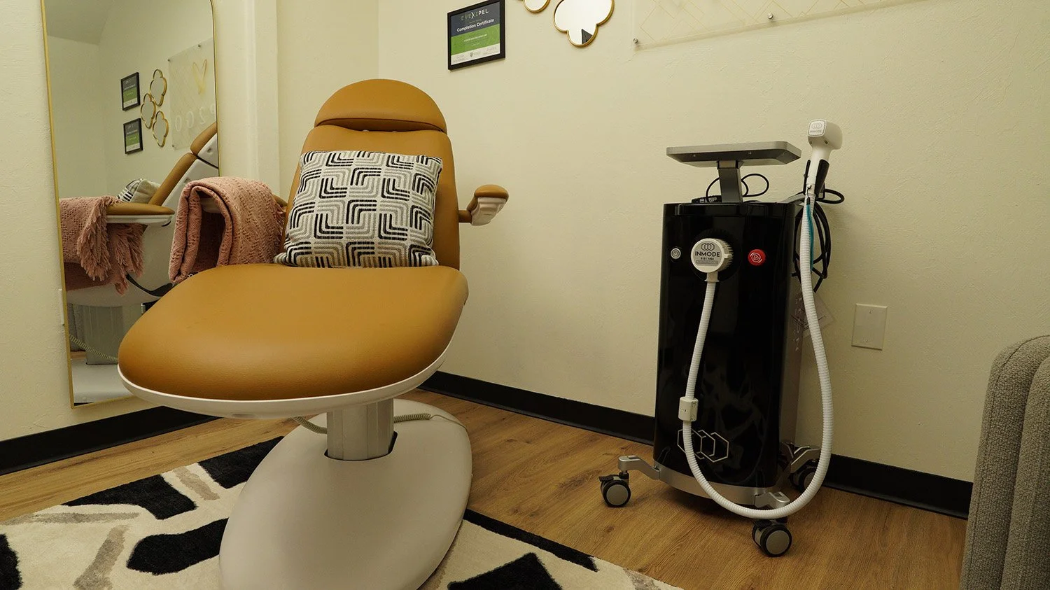 A medical or spa treatment room with a brown reclining chair, a patterned pillow, a mirror, pink towels, and medical equipment on a mobile stand.