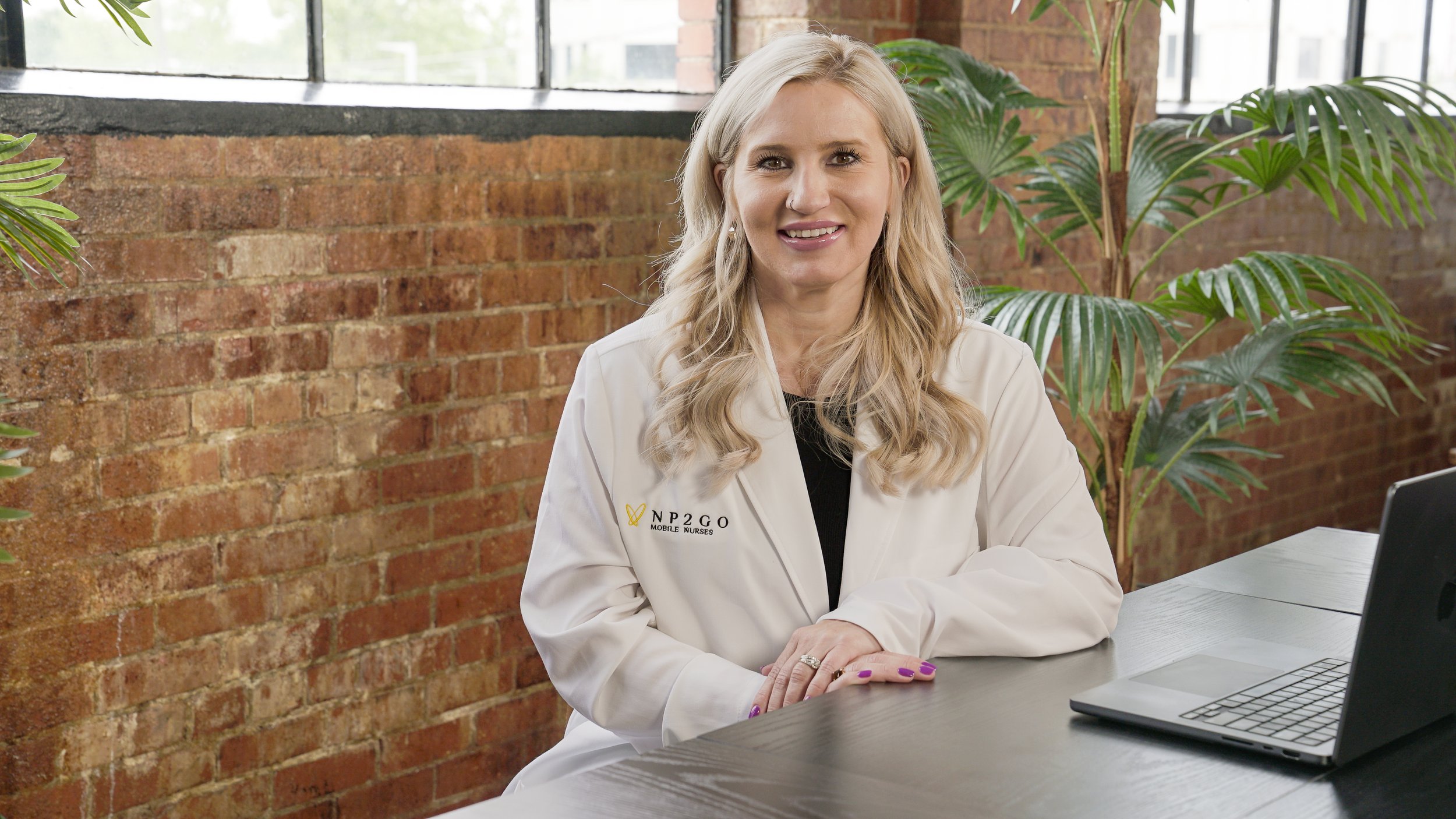 A woman with blonde hair in loose waves wearing a white jacket with a logo that reads NP2GO Mobile Nurses, sitting at a wooden table with a laptop in front of her, in a room with large windows, a brick wall, and indoor plants.