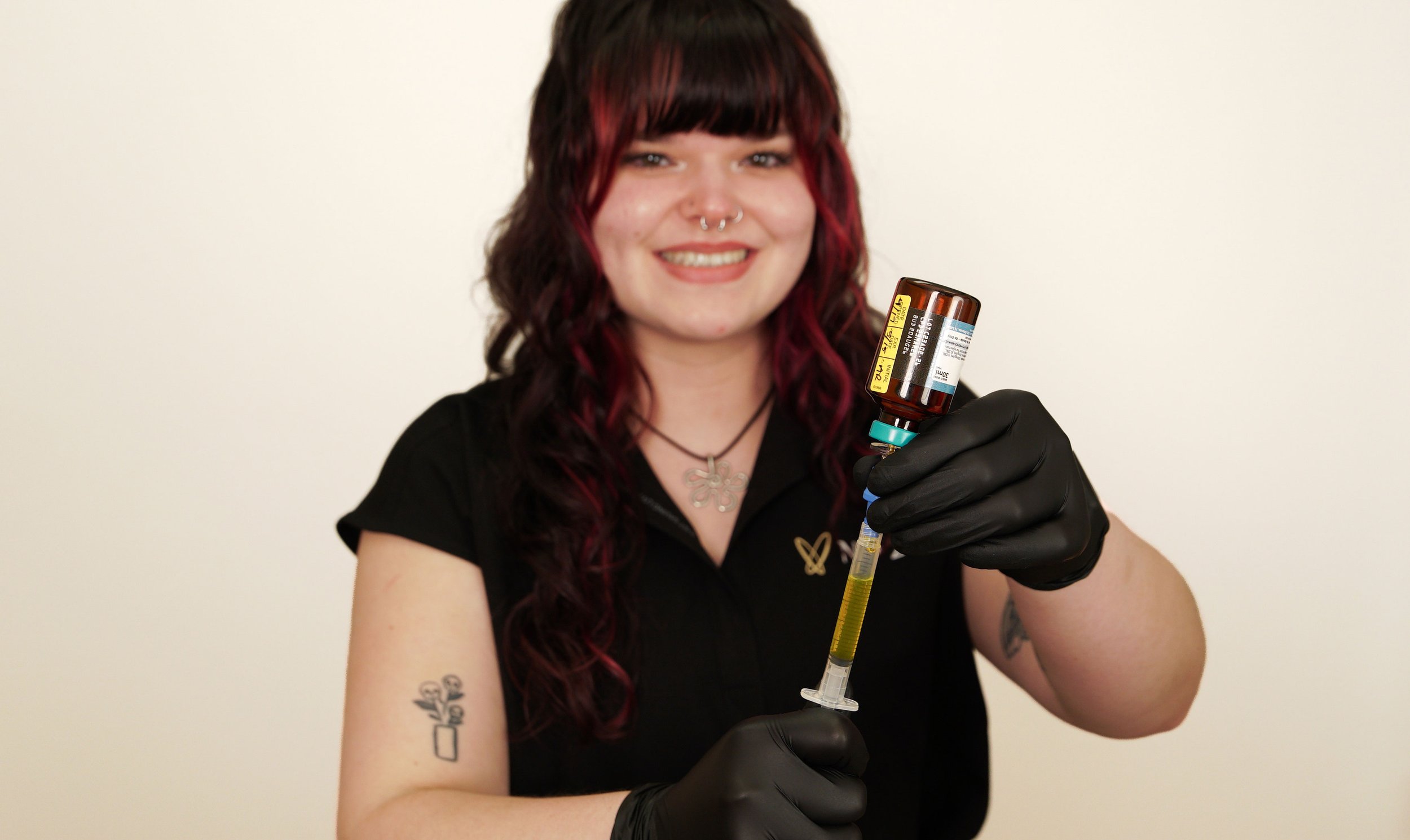A young woman with dark red hair, tattoos on her left arm, and a nose piercing holds a syringe filled with yellow liquid, smiling at the camera. She wears black gloves and a black shirt with a butterfly pendant necklace, standing against a plain white background.