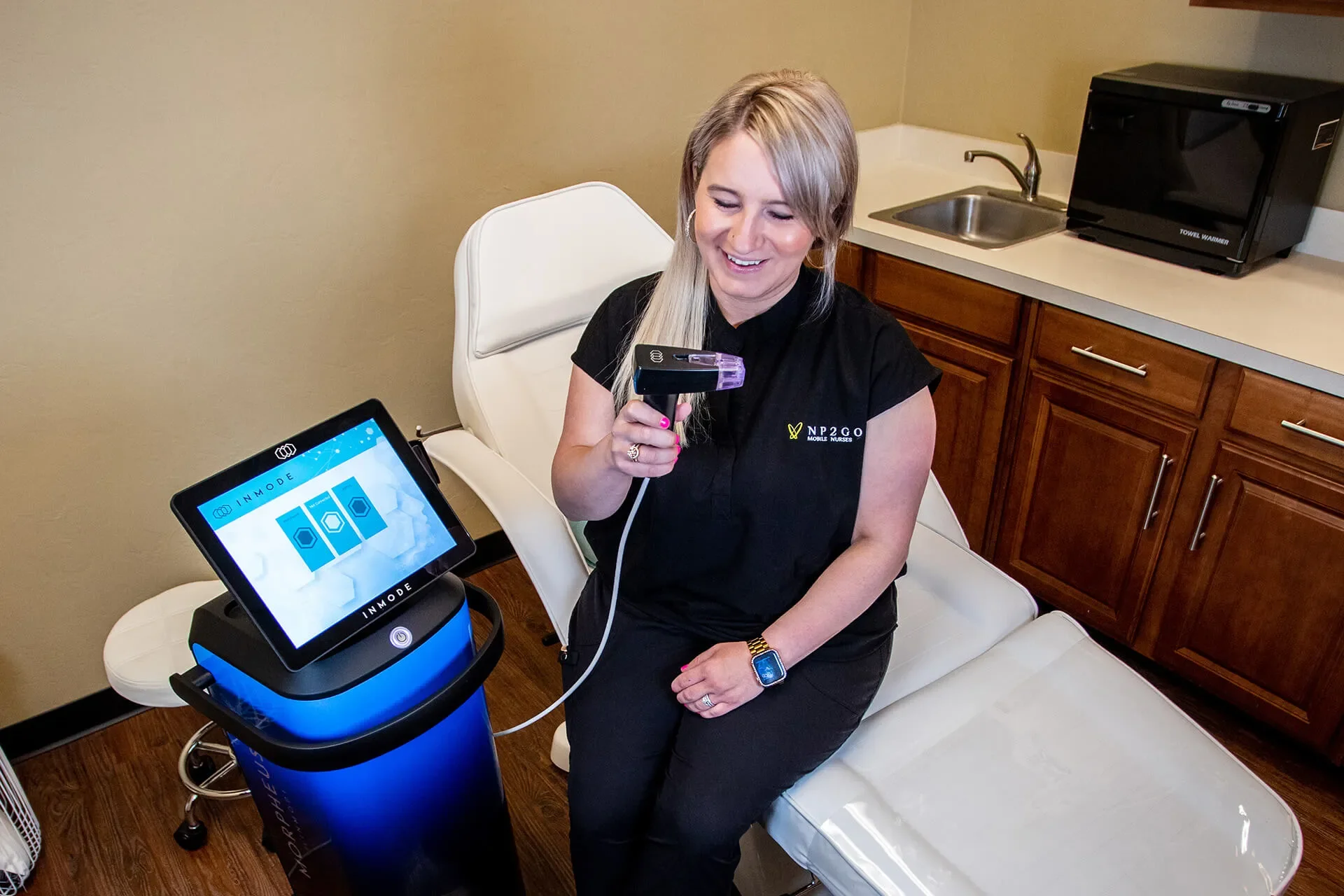 A woman with blonde hair, wearing a black shirt, sitting on a chair, smiling while using a medical device that appears to be a blood pressure monitor or similar diagnostic tool, with a screen displaying colorful graphics in the foreground.