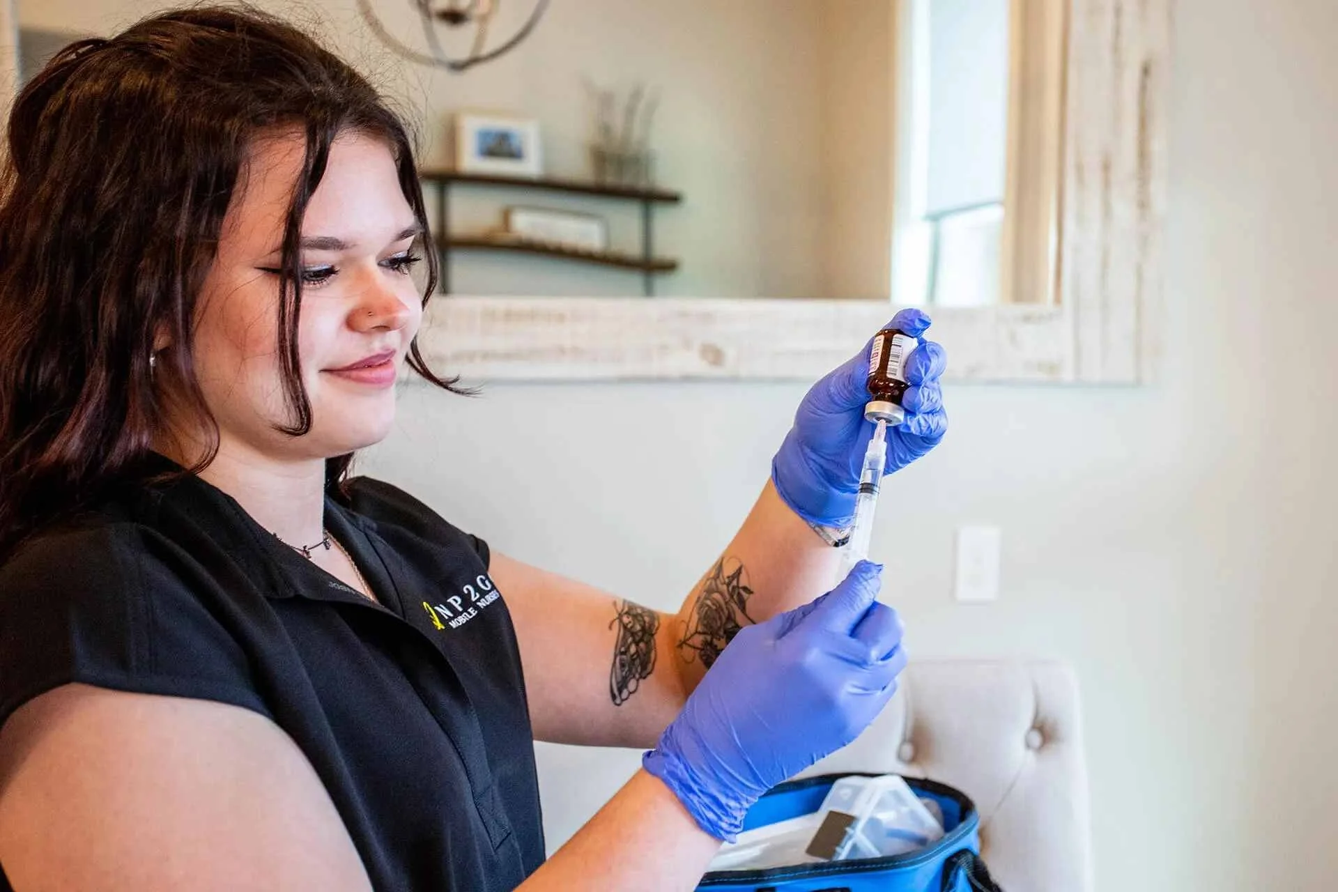A woman with dark hair wearing purple gloves is holding a syringe and preparing to give an injection with a vial. She has visible tattoos on her left arm and is wearing a black shirt.
