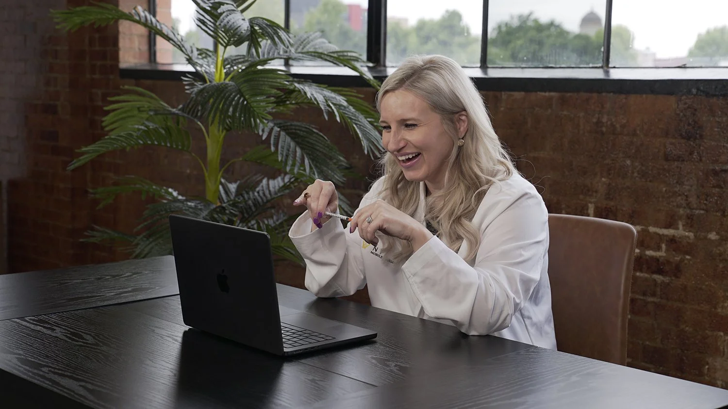 A woman with blonde hair sitting at a dark wood table in front of a laptop, smiling and holding a pen with both hands. Behind her, a large potted plant and a window showing an outdoor view.
