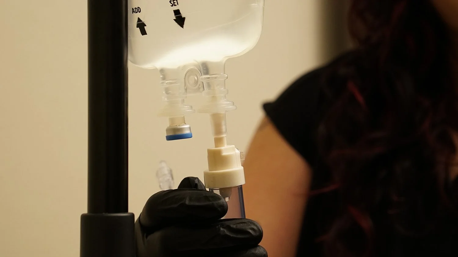 Close-up of a person receiving a vaccination or injection in their upper arm, with a healthcare professional wearing black gloves holding a syringe, and an IV bag hanging in the background.