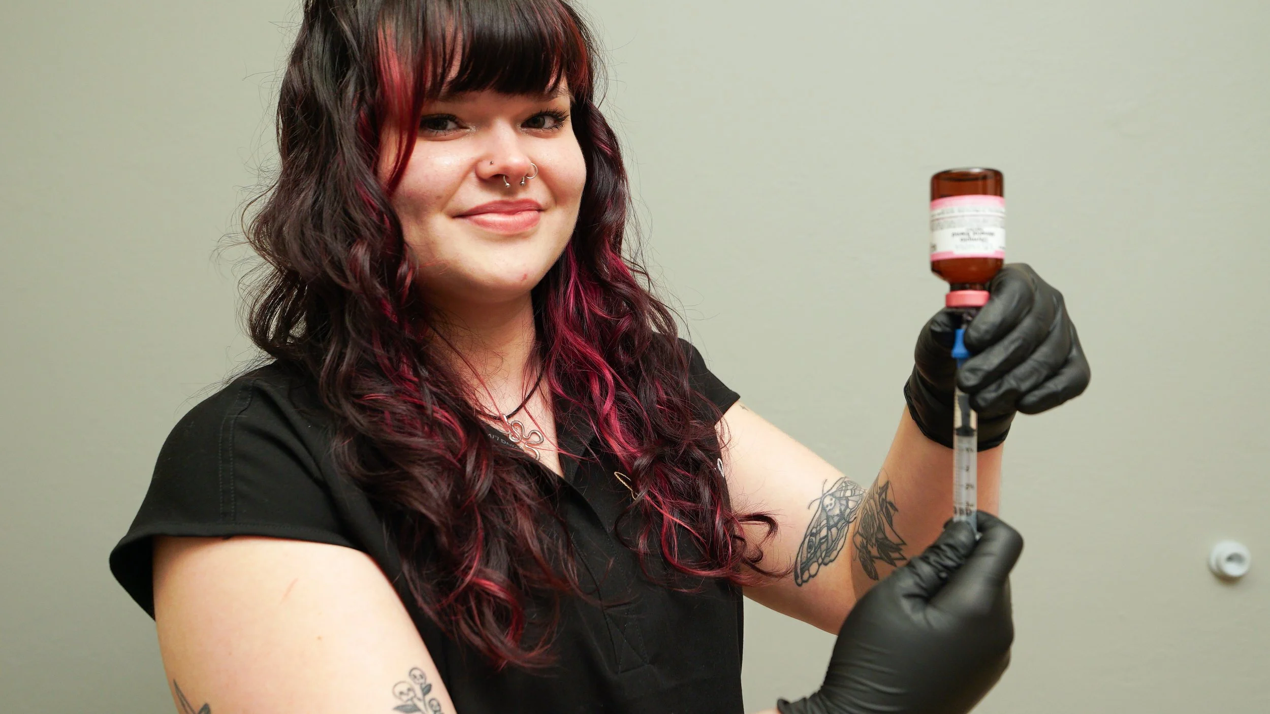 A woman with long dark hair and red highlights, wearing a black shirt, holding a medical syringe, wearing black gloves, smiling, in a plain room.