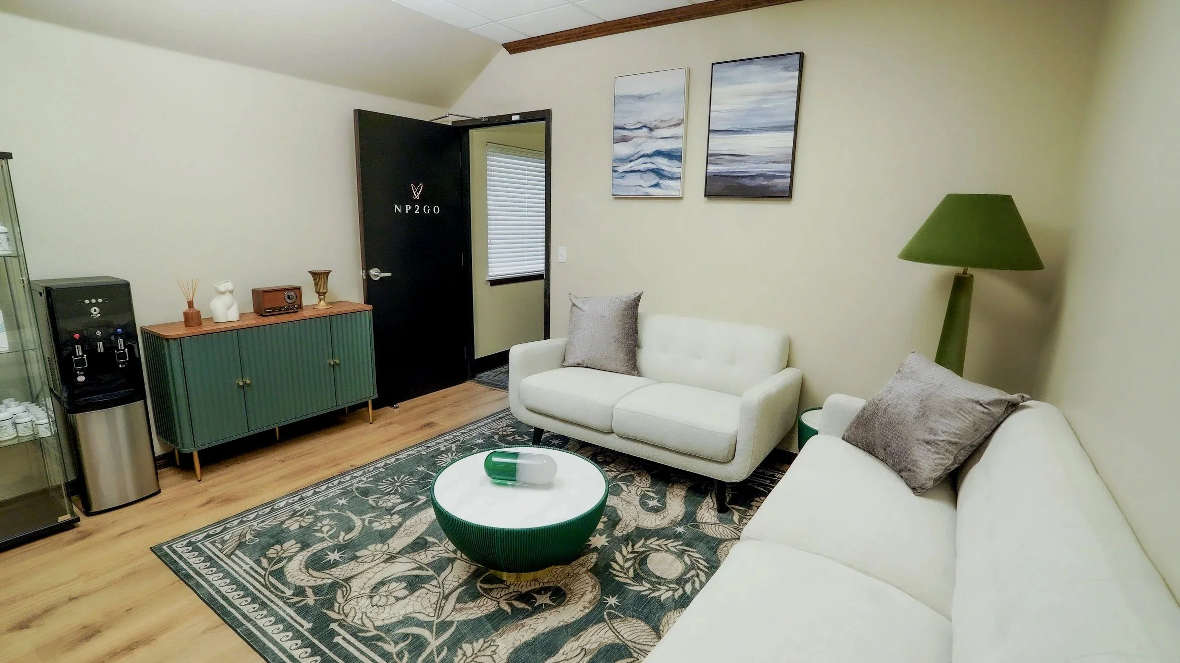 A cozy living room with white sofas, gray pillows, a green and white round coffee table, a green lamp, wall art, a cabinet, a water dispenser, and a door with labeled writing.