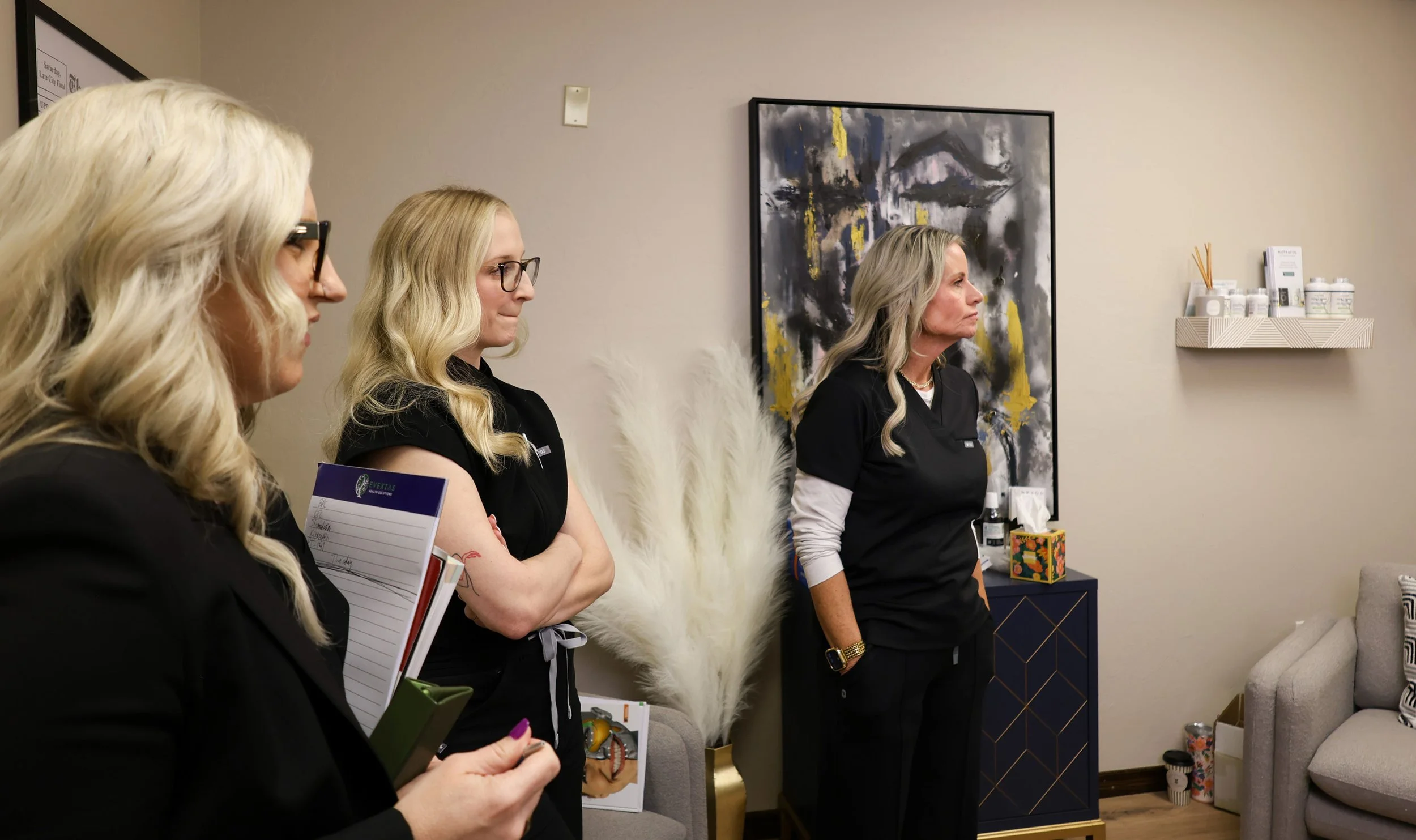 Four women with blonde hair standing in a room, listening attentively. They are wearing black tops, with notebooks and pens. Behind them, there is a large abstract painting, a white shelf with bottles and reed diffusers, and a corner of a beige sofa with a white fluffy chair nearby.