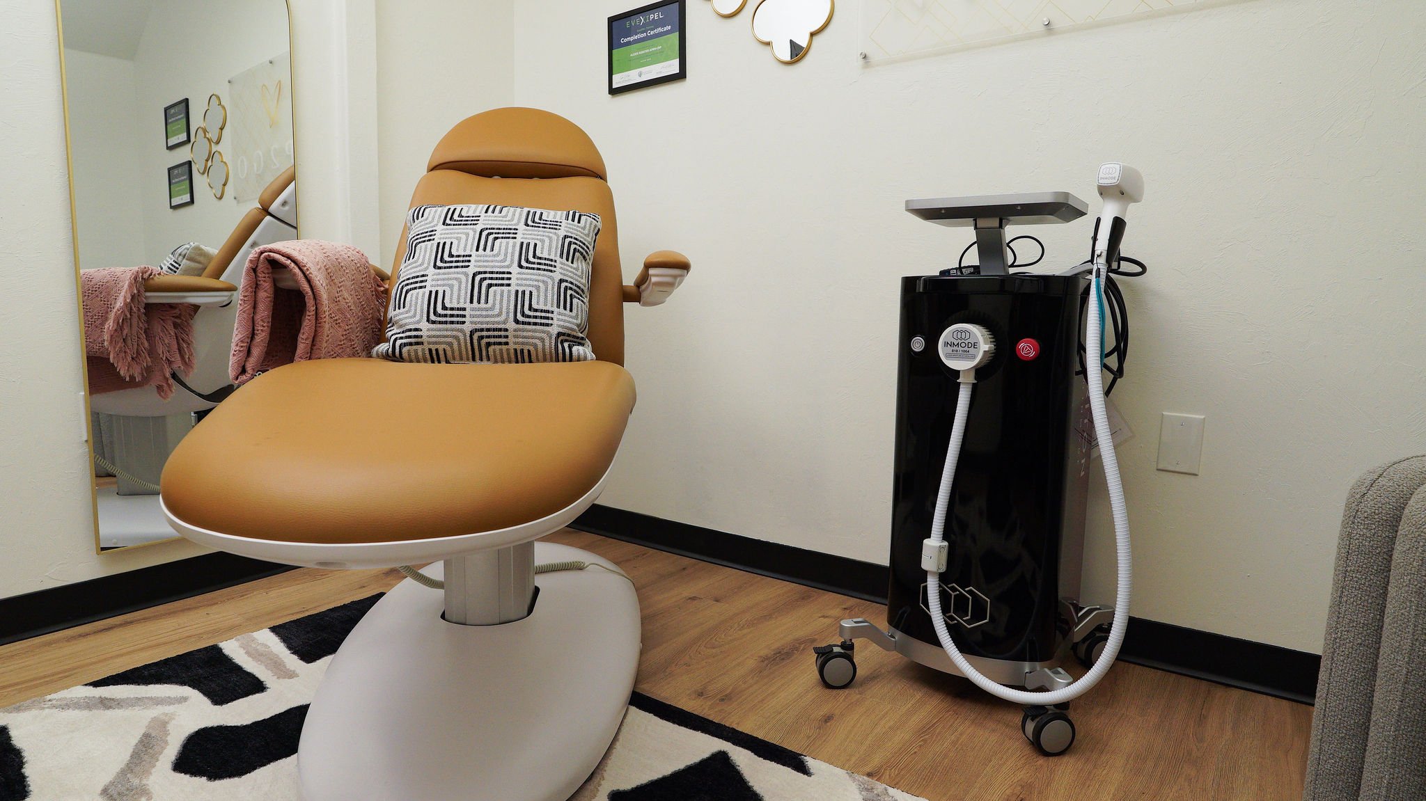 A medical or aesthetic treatment room featuring an ergonomic chair with a geometric-patterned pillow, a pink blanket, a wall-mounted mirror, framed certificates, and a medical device on a wheeled stand with tubing and handheld tools.