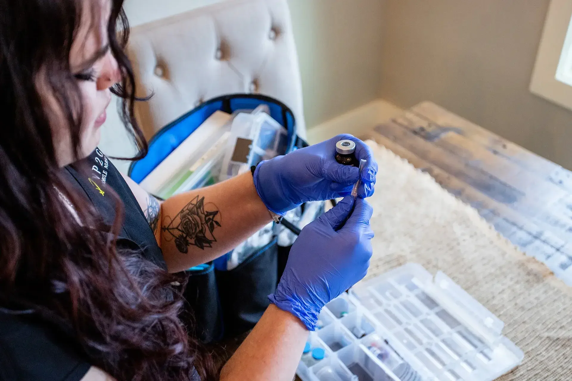 A woman with long curly hair and a tattoo on her forearm, wearing purple gloves, prepares a medical or cosmetic device with a small circular tip. She is standing at a table with a plastic organizer containing different supplies in a room with neutral-colored walls and a window.