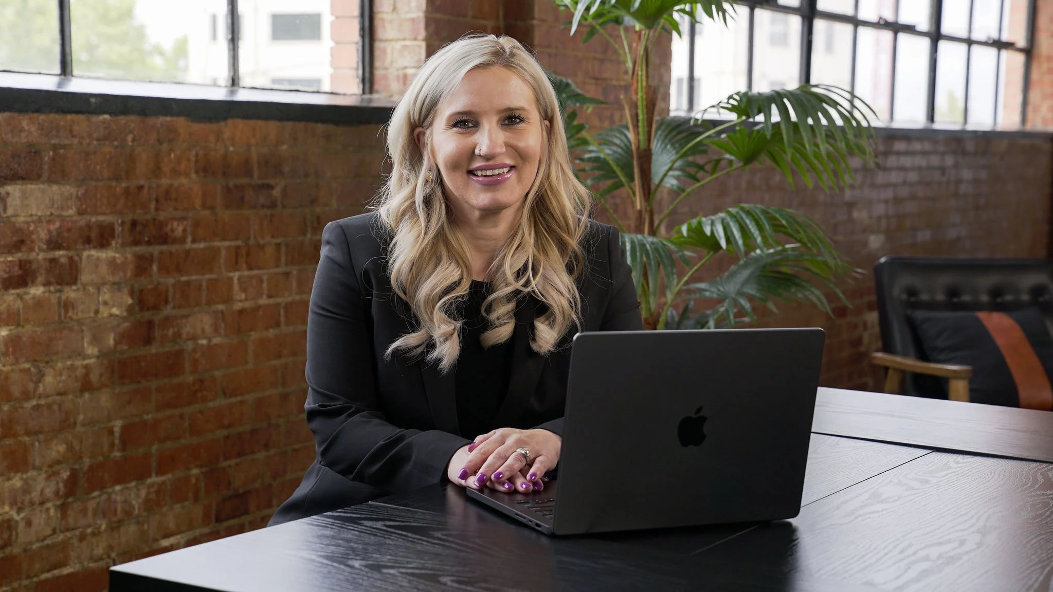A woman with long blonde wavy hair, wearing a black blazer, sitting at a desk with a laptop, smiling at the camera in a modern office with brick walls and large windows, with green plants in the background.