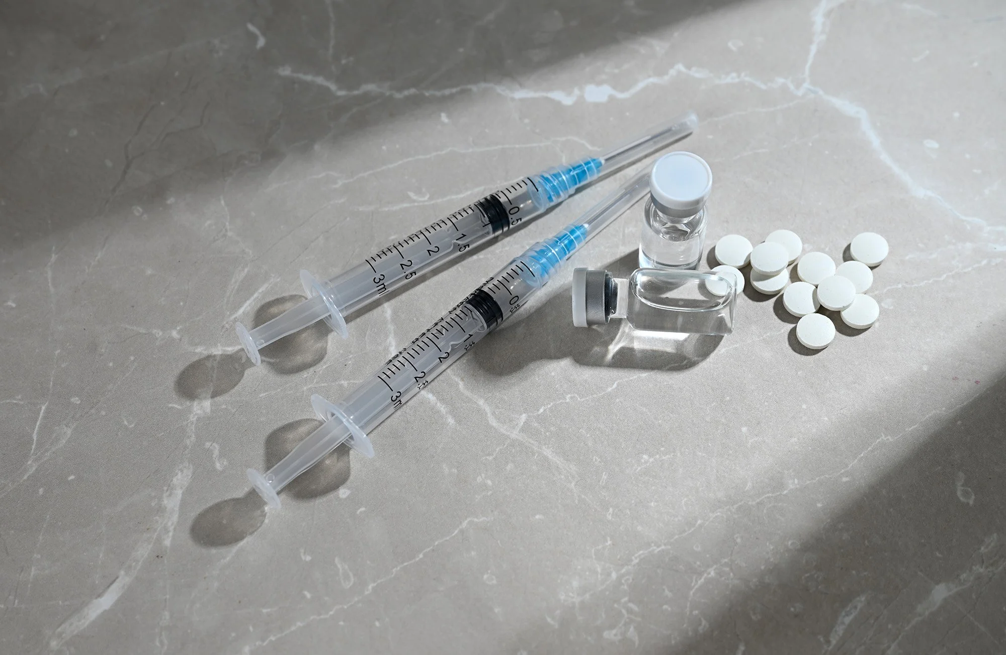 Two medical syringes, an ampoule, a small vial with a blue cap, round white tablets, and a small empty glass container on a marble surface.