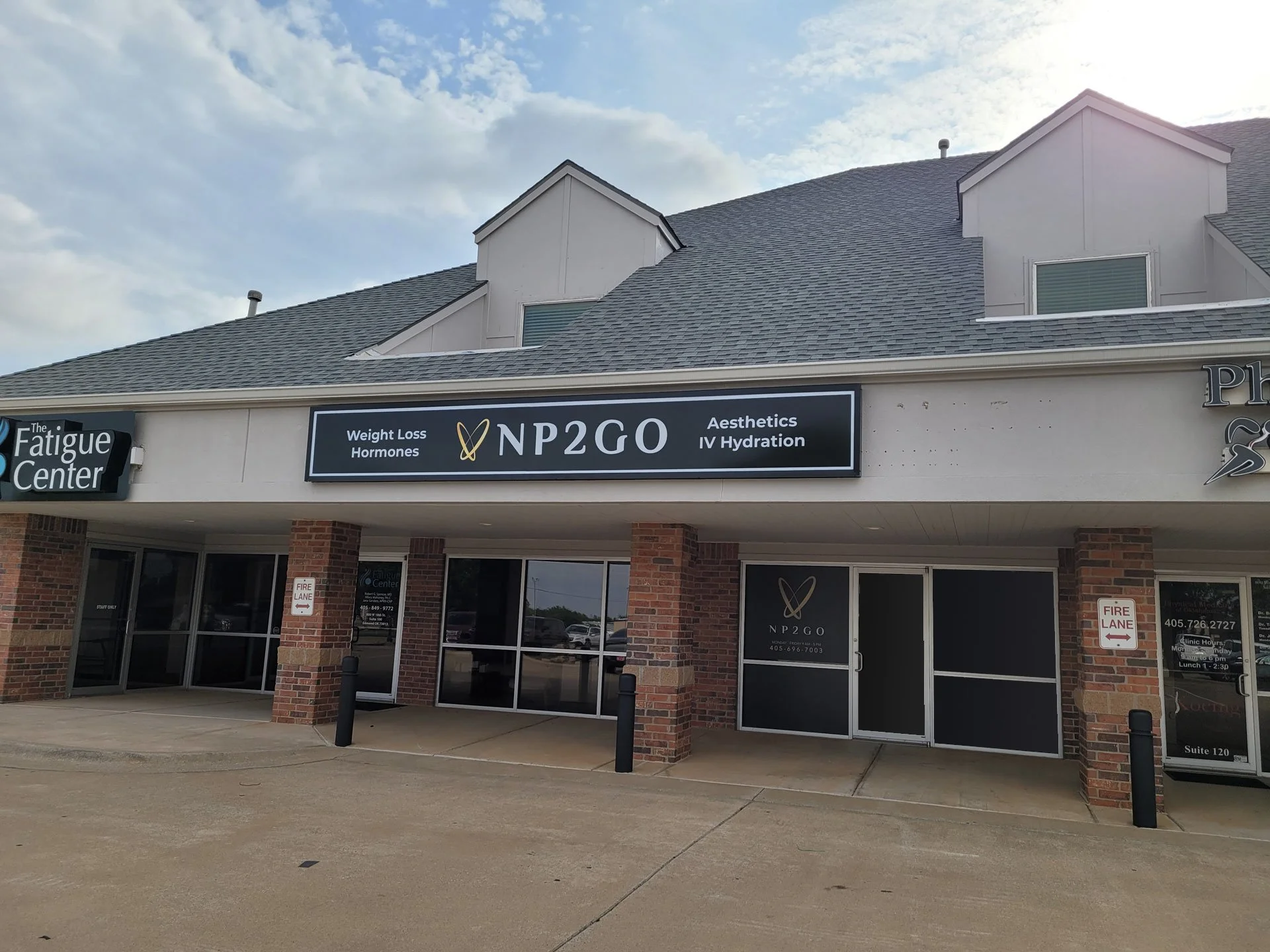 Front view of a commercial building housing a non-profit organization called NP2GO and a fatigue center. The building has brick columns, glass doors, and signage indicating services like weight loss, hormones, aesthetics, and IV hydration. The parking lot is empty with a concrete sidewalk, and the sky above is partly cloudy.
