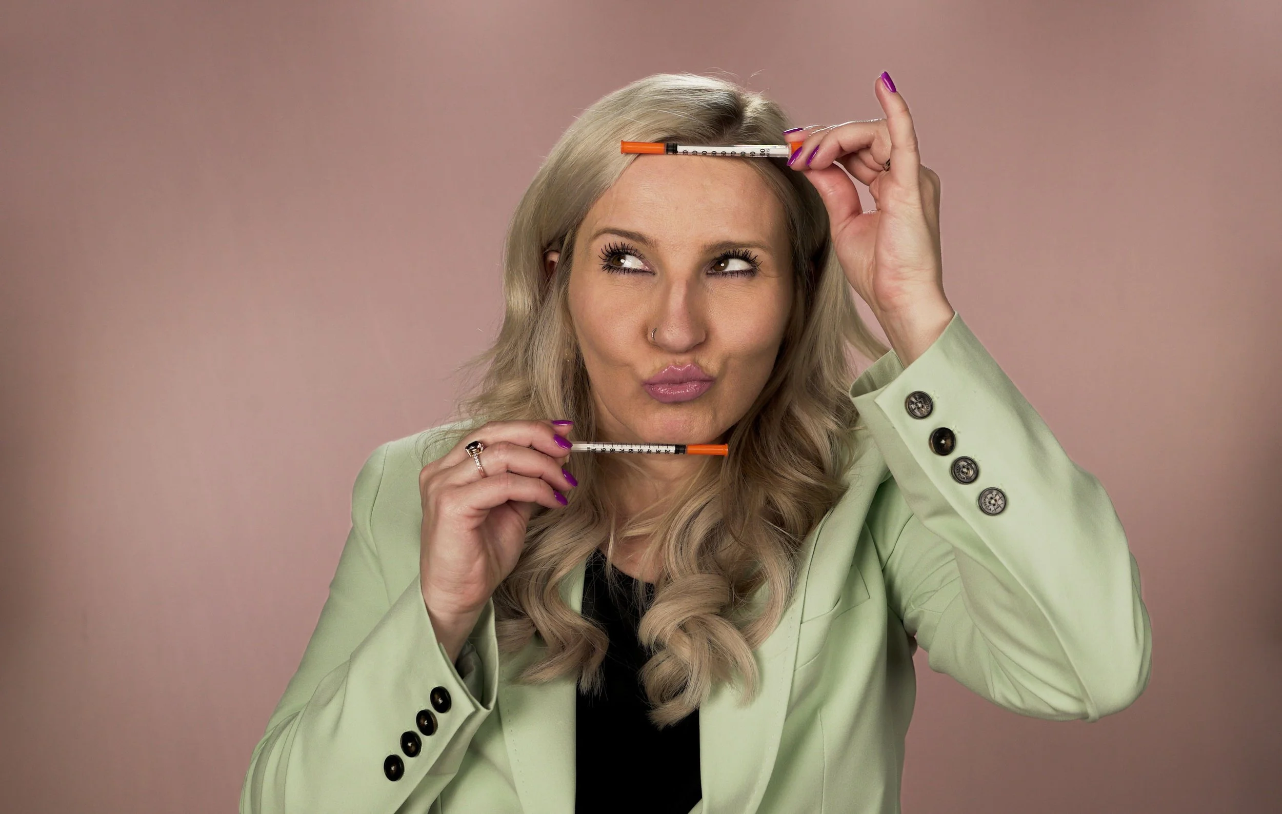 A woman with long blonde hair is holding two syringes, one above her head and one below her chin, while looking to the side with a playful expression, wearing a pale green blazer and a black top against a pink background.