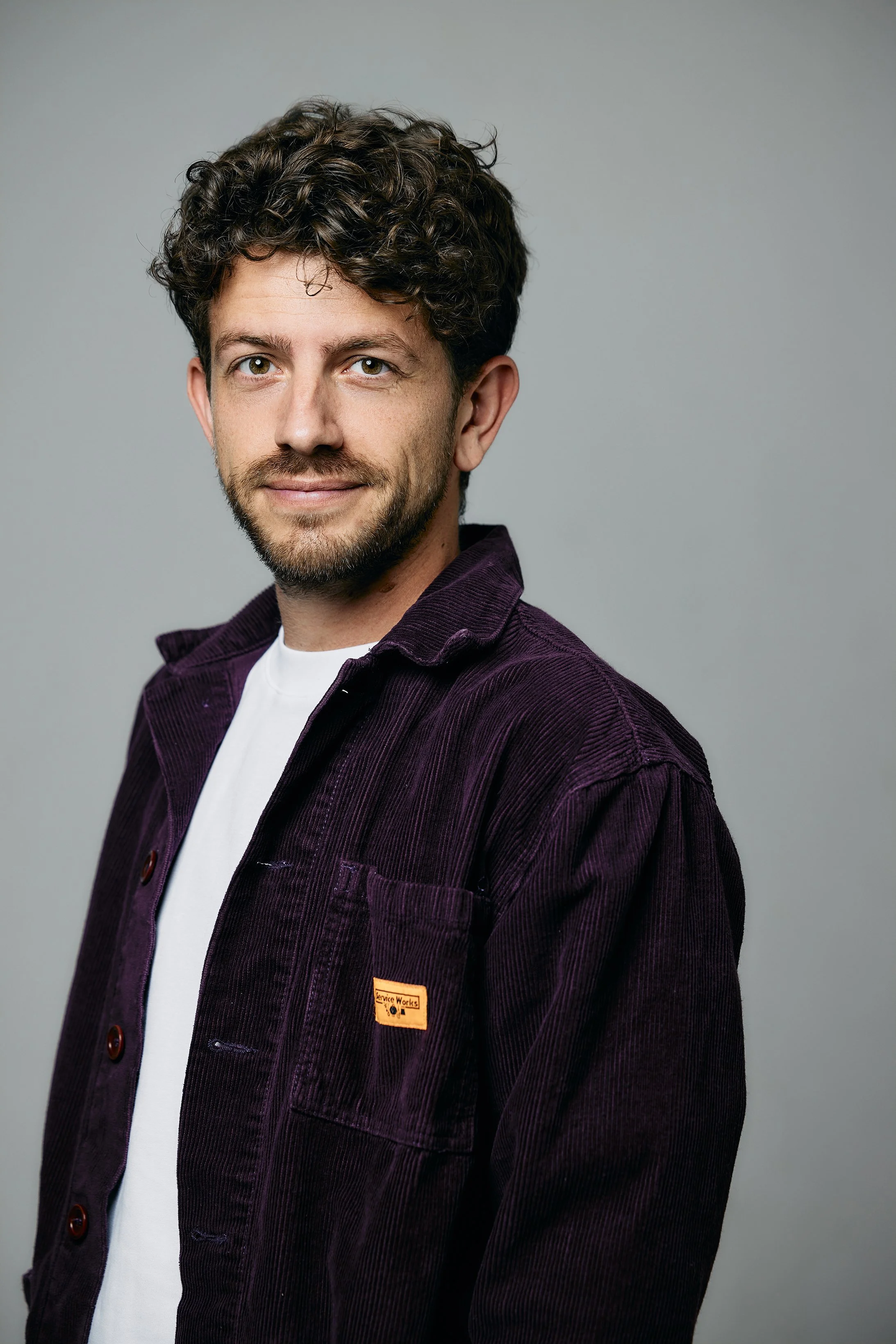 A man with curly dark hair and a beard is wearing a white shirt and a purple corduroy jacket, posing against a plain gray background.