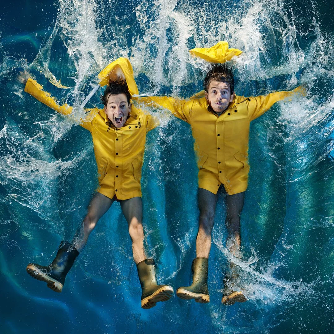 Two people in yellow raincoats and boots floating in a splash of water, looking excited and surprised.