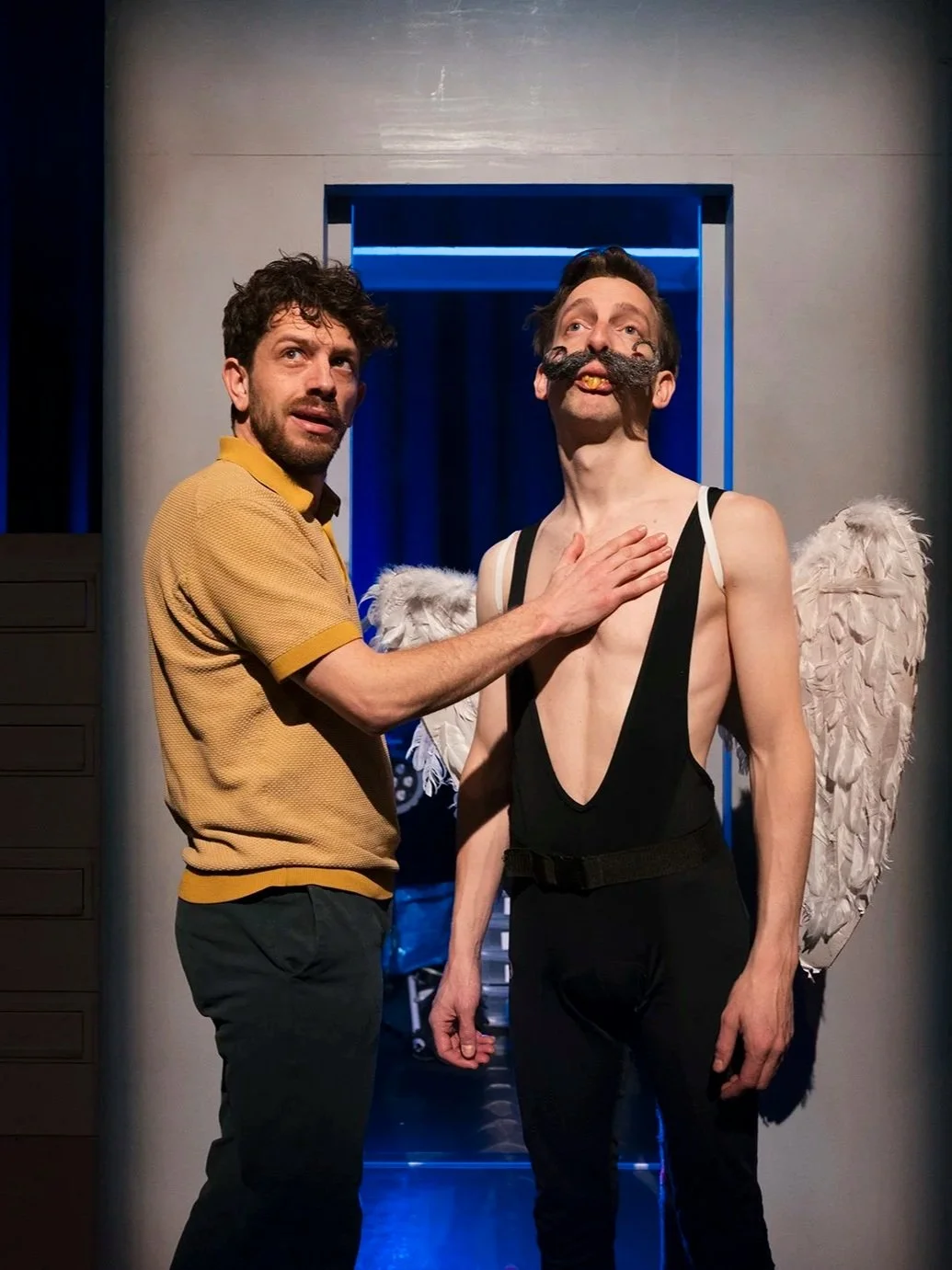 Two men on stage with a gray wall and a blue doorway in the background. The man on the right is shirtless, has a large fake mustache, angel wings, and is wearing black suspenders. The man on the left is touching the chest of the shirtless man.