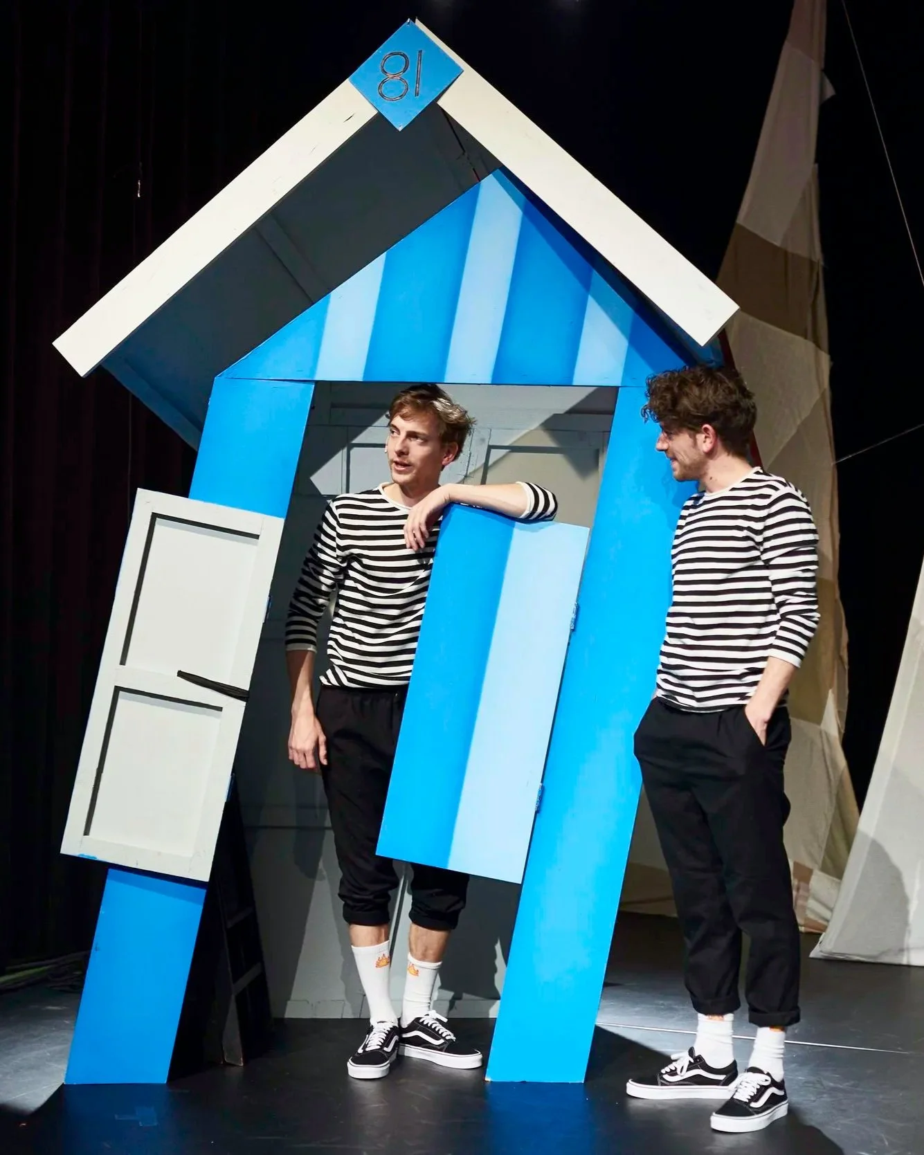 Two young men in striped shirts and black pants standing in front of a blue and white stage set resembling a house. One is leaning on a blue structure, the other has his hands in his pockets, both wearing Vans sneakers and white socks.