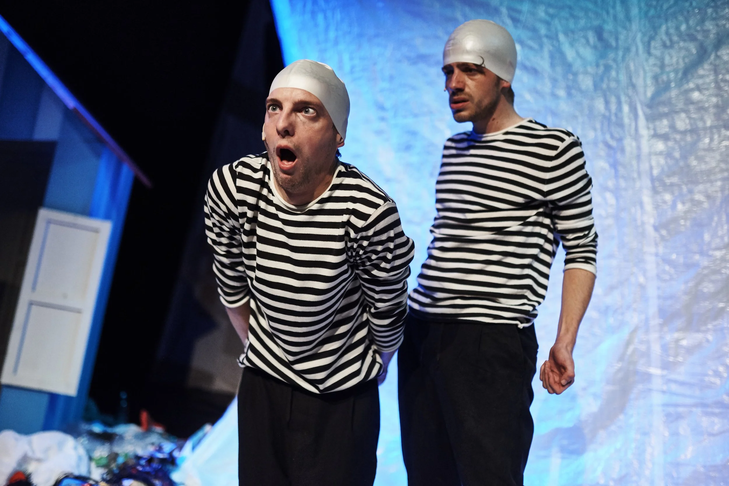 Two actors in black and white striped shirts and white head coverings performing on stage, with one actor looking surprised and the other appearing concerned.